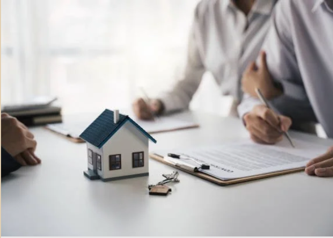 Real estate agent and clients signing documents during house sale, with a small house model and keys on the table.