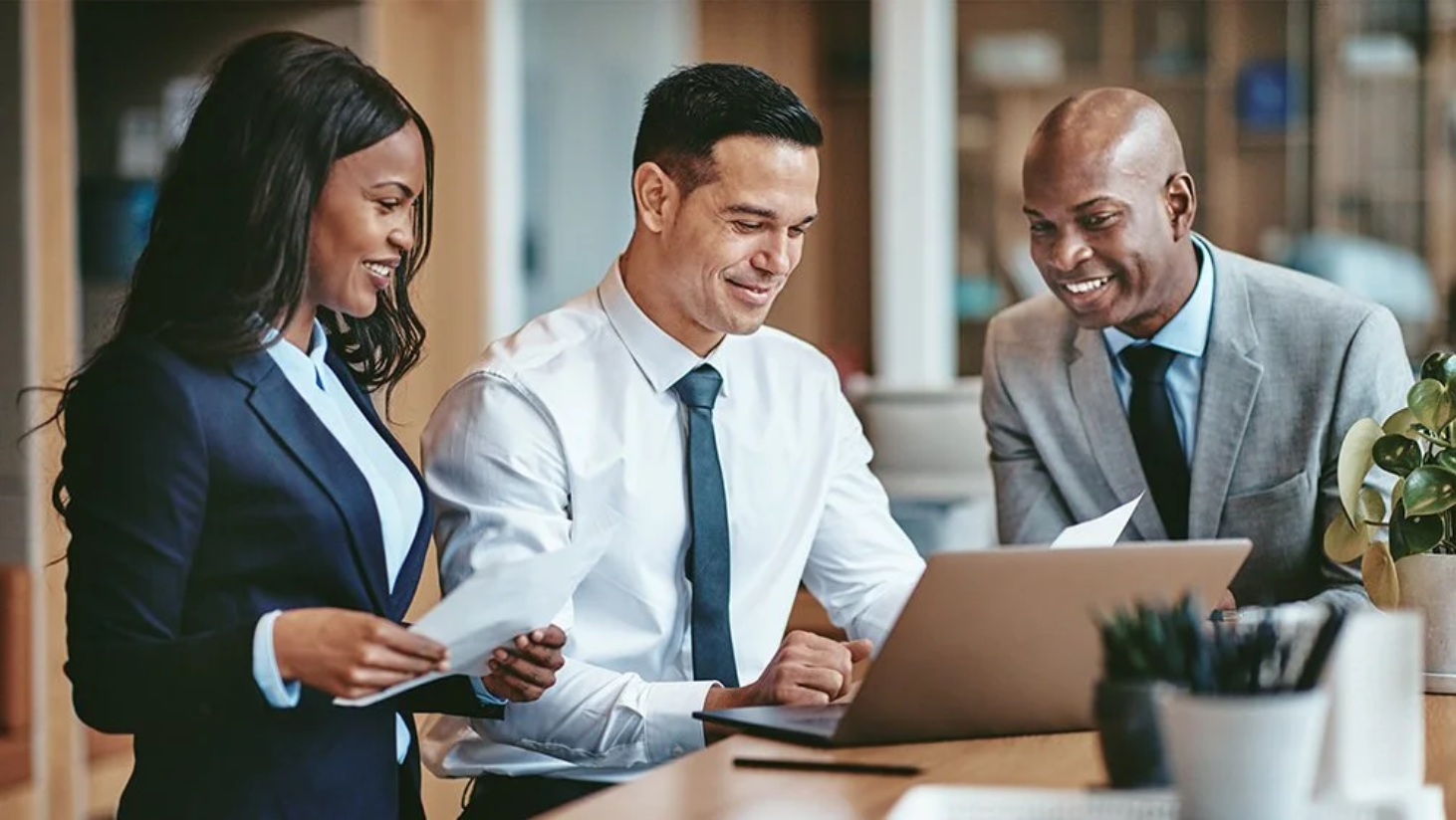 Three diverse professionals in business attire collaborating around a laptop and documents in an office.