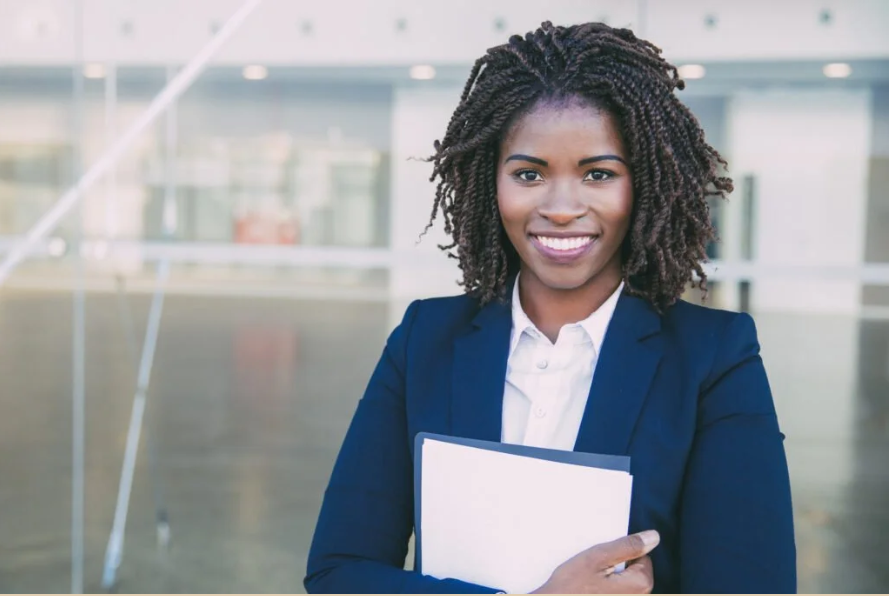 A smiling woman in a navy blue blazer holding a folder in an office building conference area.