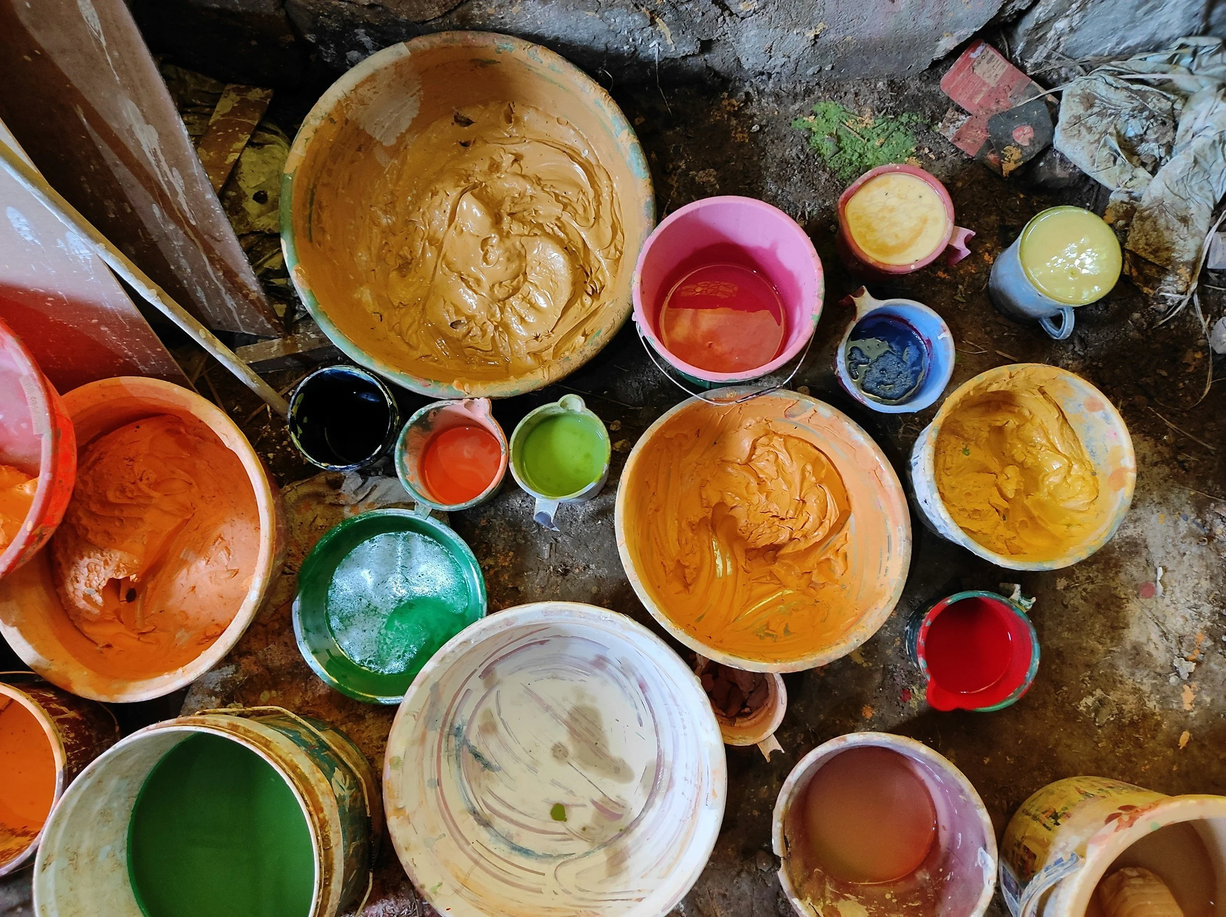 Several open paint buckets and smaller cups filled with various bright colors of paint, such as orange, yellow, green, blue, red, and black, scattered on a rough ground surface.  It depicts failure to decorate in a dilapidations context.