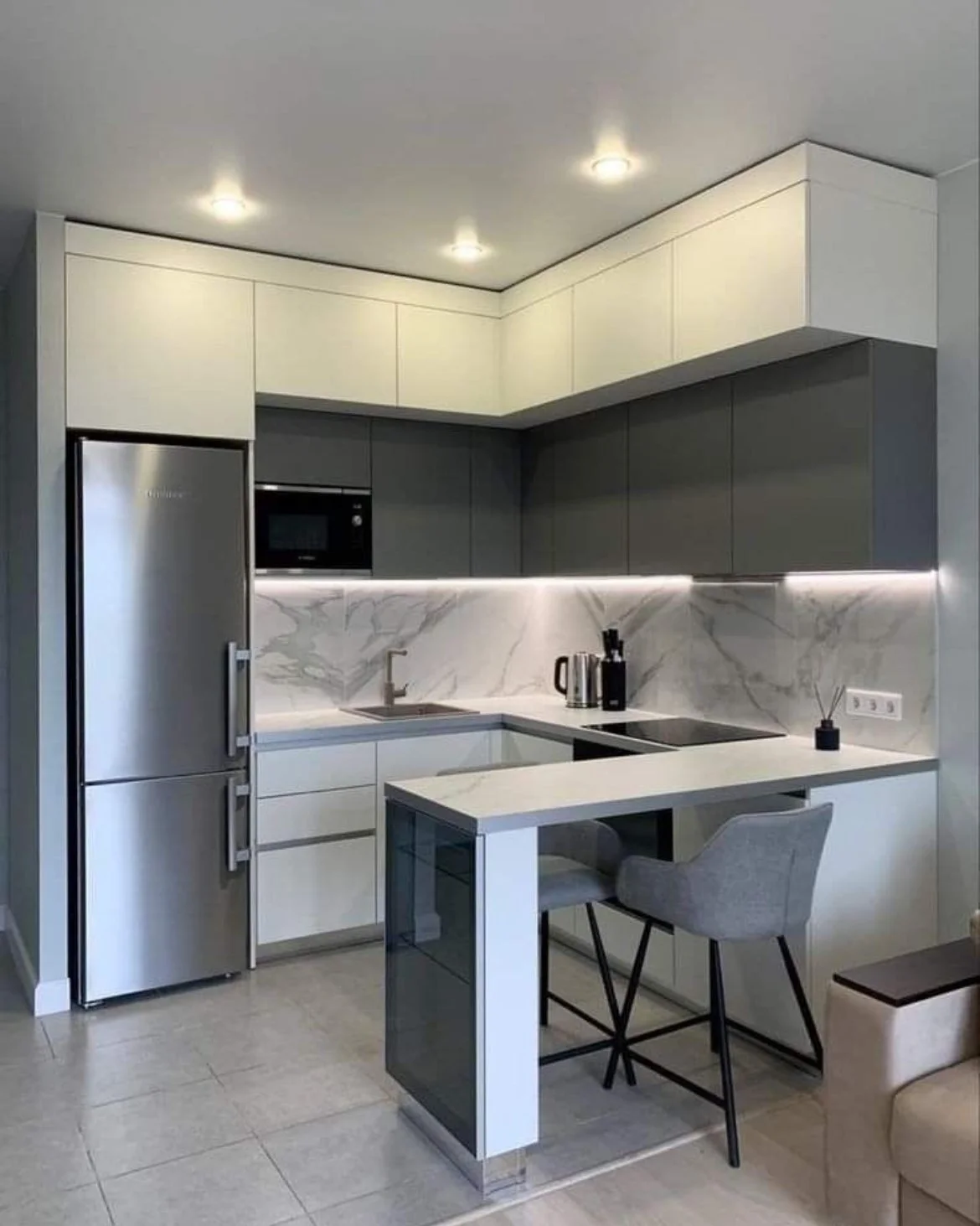 Modern kitchen with white and gray cabinets, marble backsplash, stainless steel refrigerator, microwave, black stove, and a small breakfast bar with a gray chair.