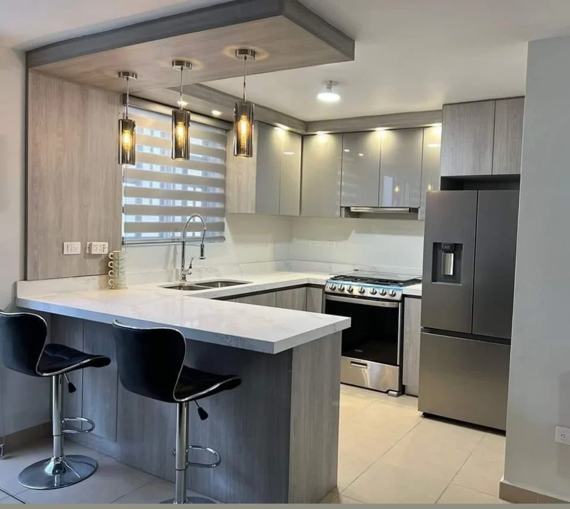 Modern kitchen with a meson blanco, gray cabinets, stainless steel appliances, and black bar stools, illuminated by pendant lights and recessed ceiling lights.