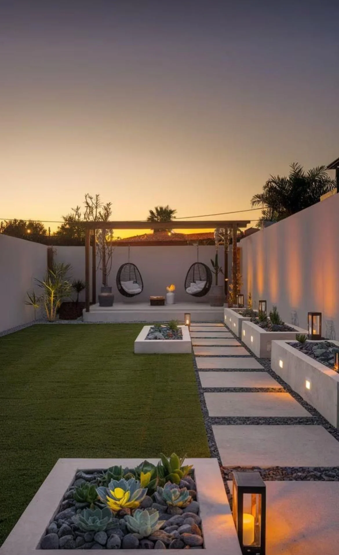 Modern backyard at sunset with a white pathway, green lawn, potted succulents, and a sitting area with hanging chairs and cushions surrounded by warm lighting.