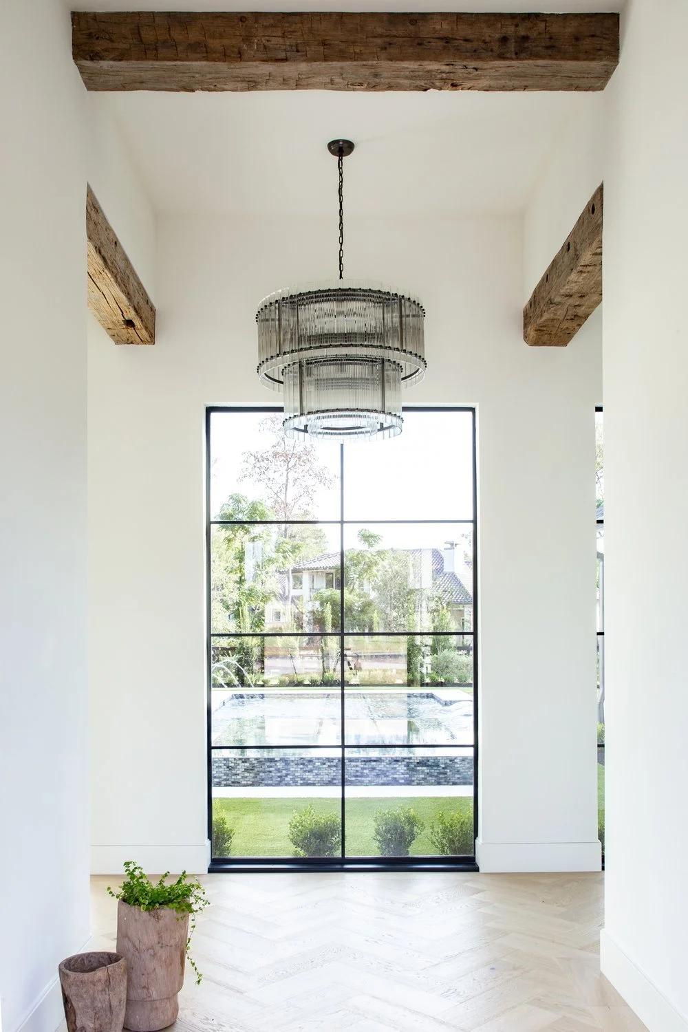 Interior view of a modern space with a large window showing a backyard with a pool, wooden ceiling beams, a chandelier hanging from the ceiling, and two pots with plants on the floor.