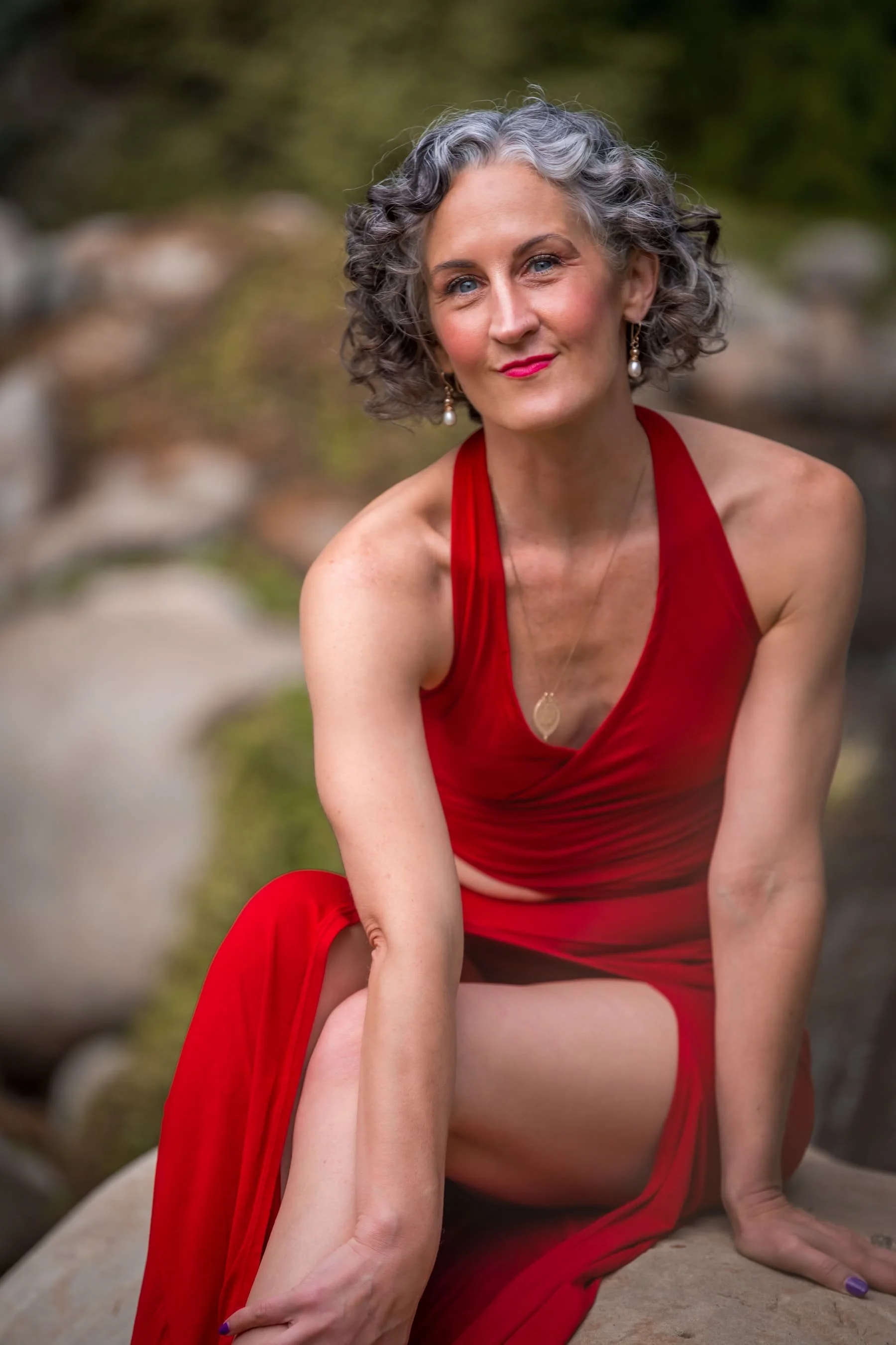 A mature woman with curly gray hair, wearing a red dress, earrings, and a necklace, sitting outdoors on rocks with a blurred natural background.