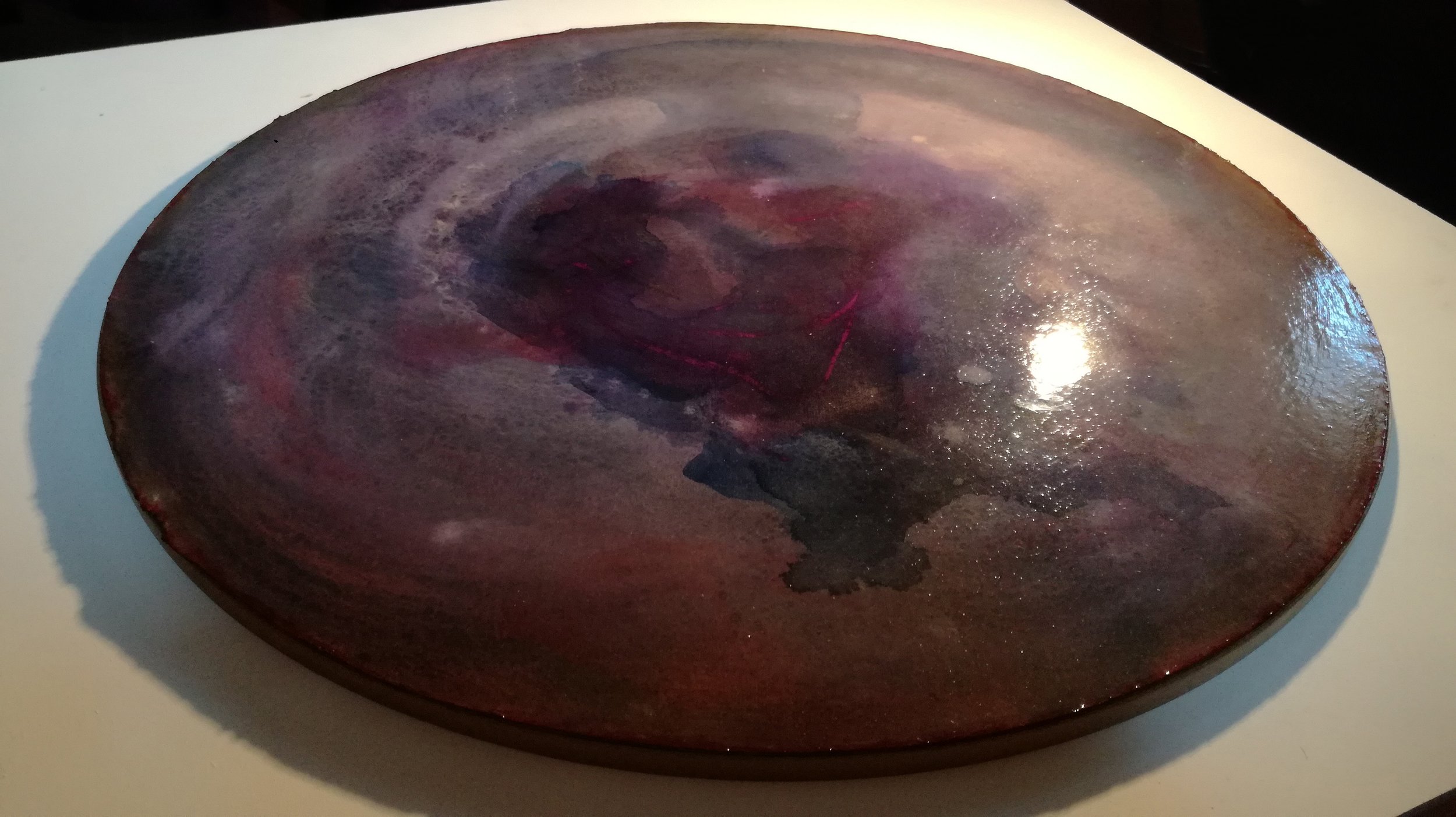 A round, flat, marbled-looking object with swirling dark red, purple, and black colors, placed on a white surface.