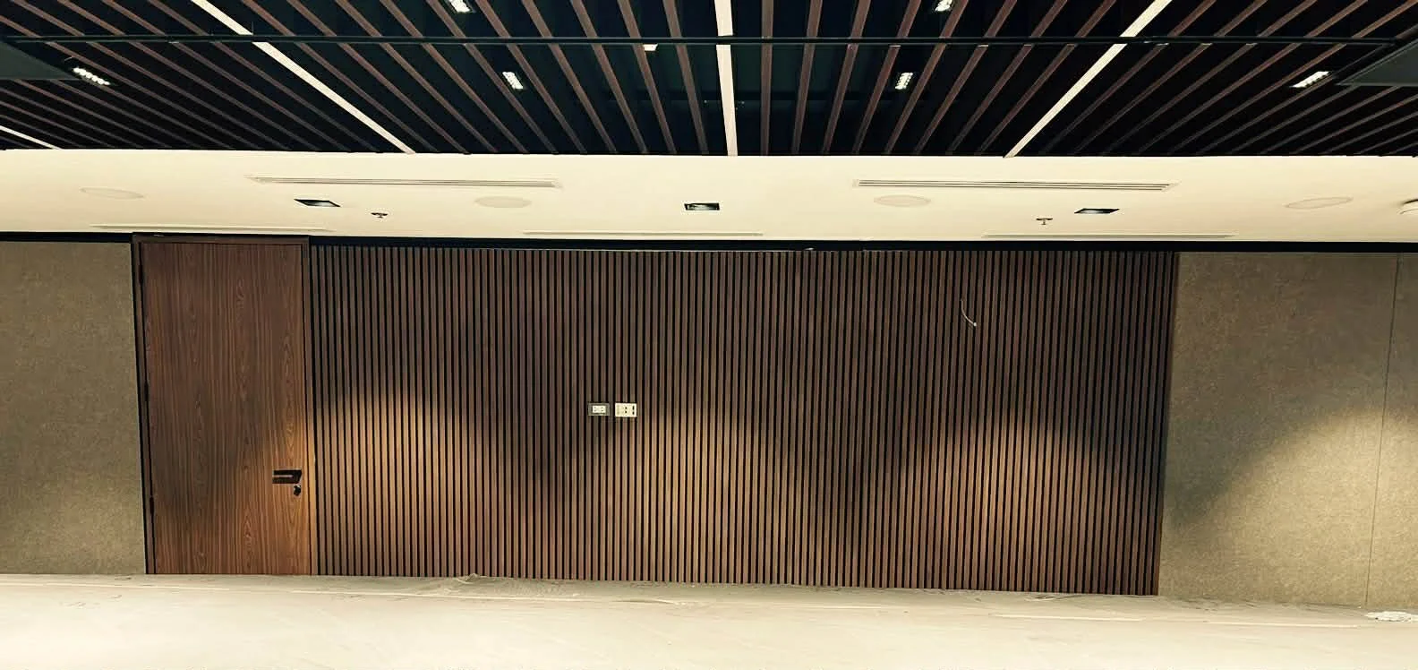 Empty hallway with wood-paneled wall, electrical outlets, and ceiling with rectangular light fixtures.