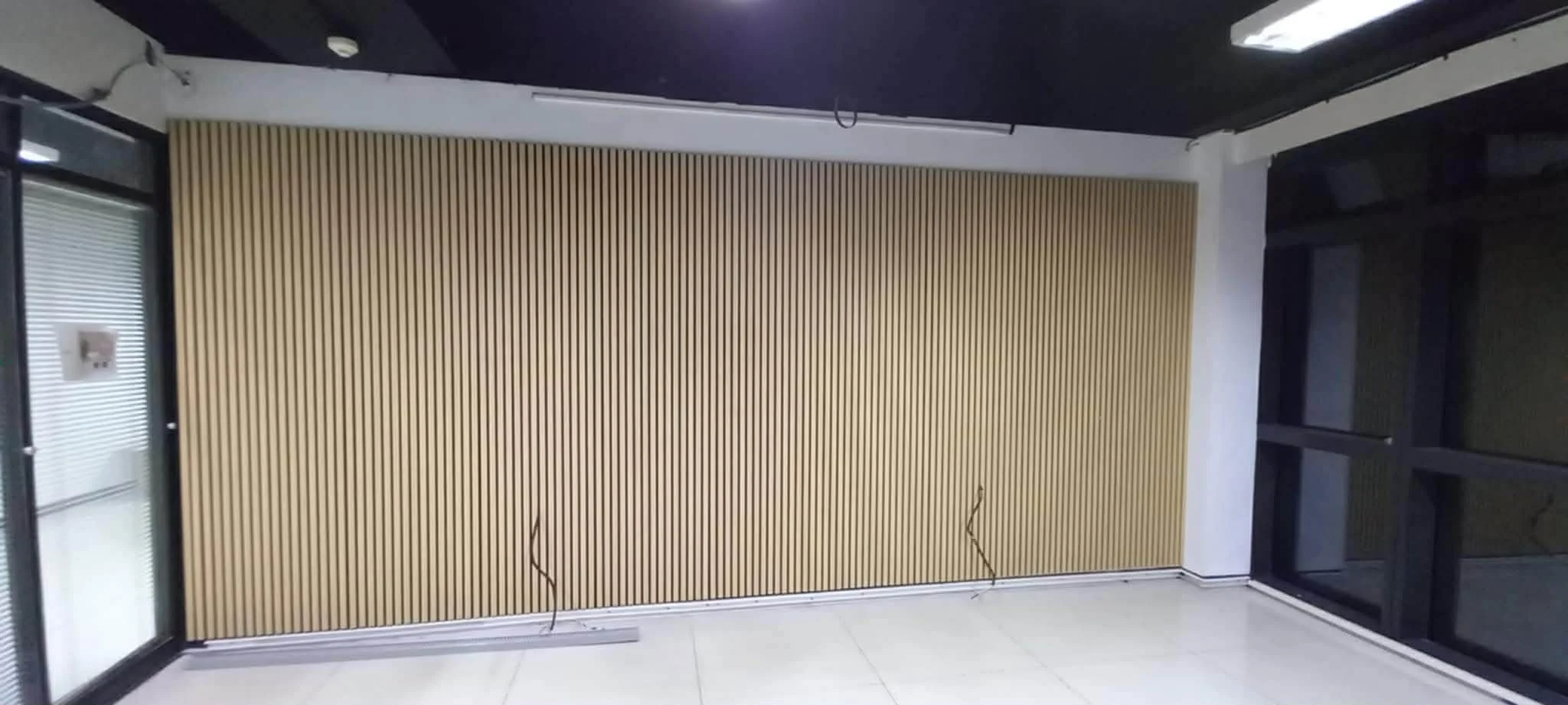 Interior of a room with a large wooden slat wall, sliding glass door on the left, and large window on the right, with black framing and ceiling lights.