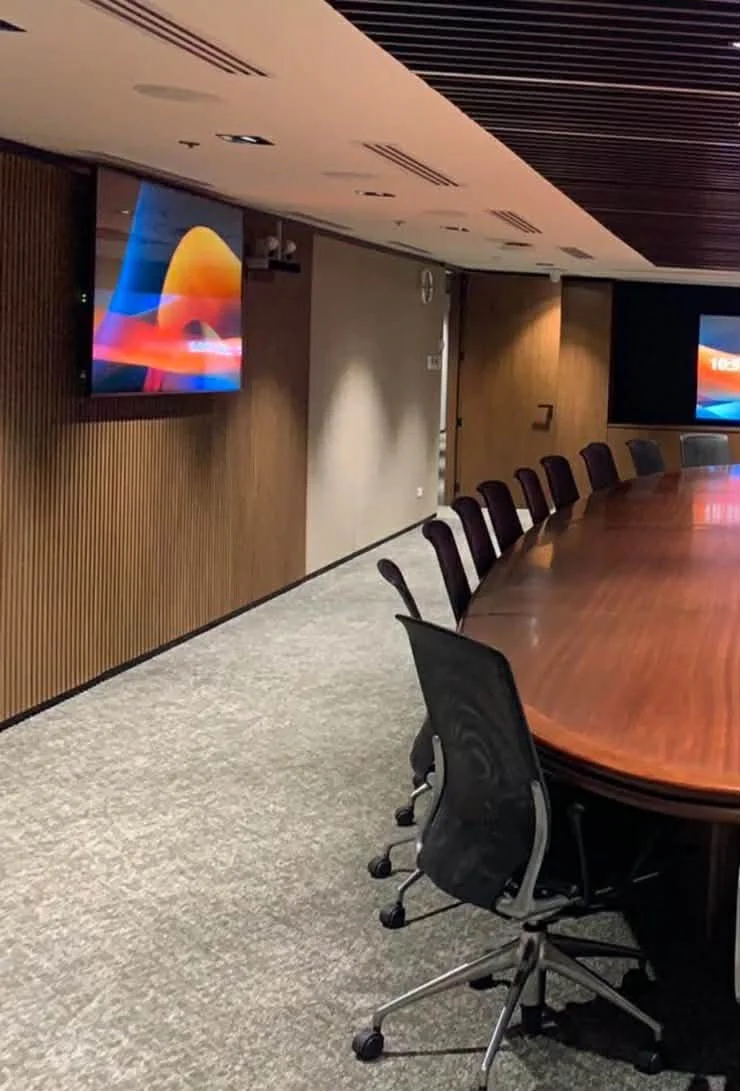 Empty conference room with a large oval wooden table and black chairs. Two wall-mounted flat-screen TVs displaying colorful abstract images. Modern ceiling design with wood paneling and air vents.