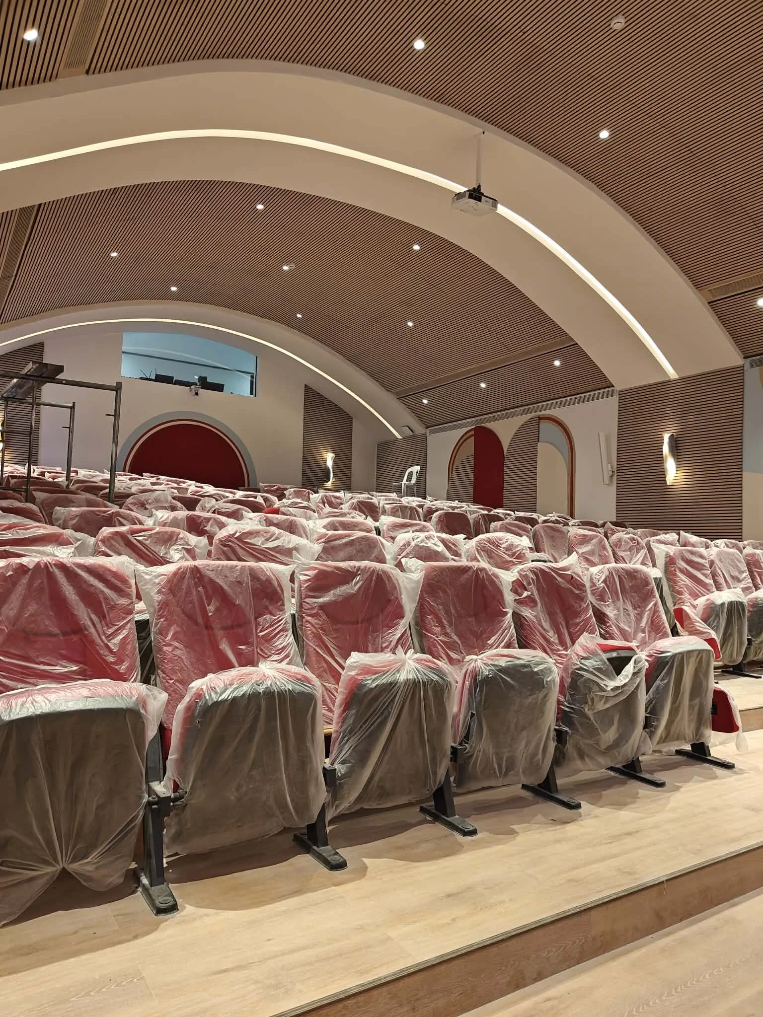 Empty auditorium with red cushioned seats covered in plastic, wooden floors, arched ceiling with recessed lighting, and a projector hanging from the ceiling.