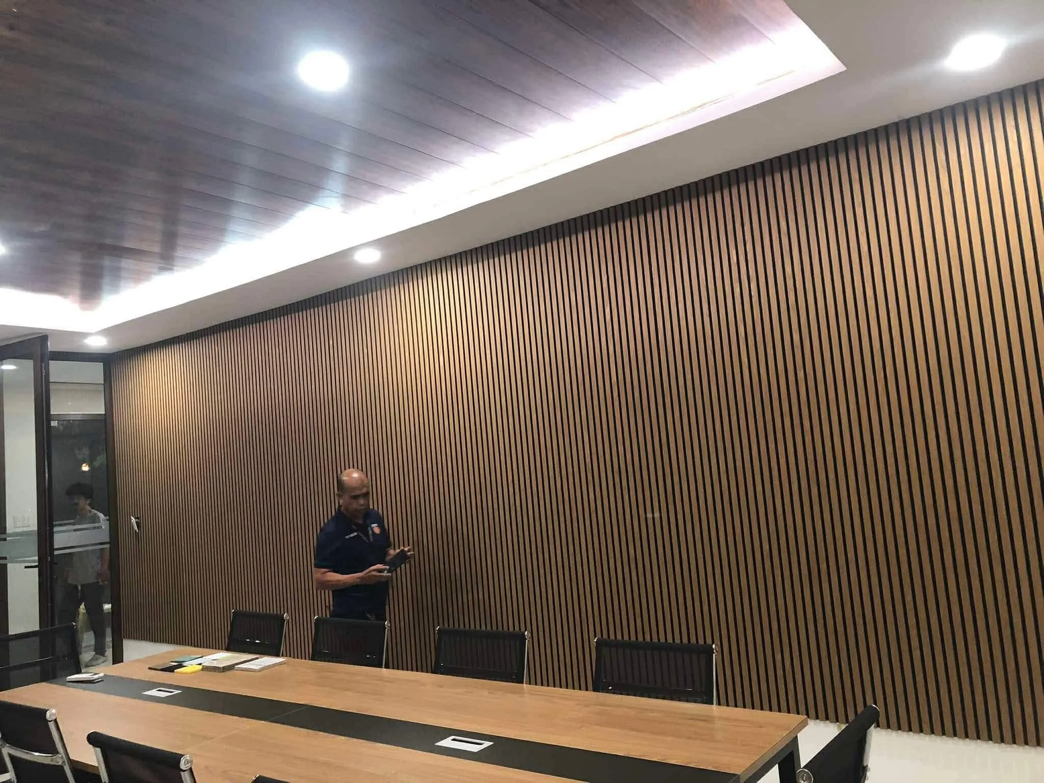 A modern conference room with a long wooden table and black chairs. There is a man standing near a large wooden wall with vertical slats, looking at his phone. The room has a ceiling with recessed lighting and a glass door on the left side.