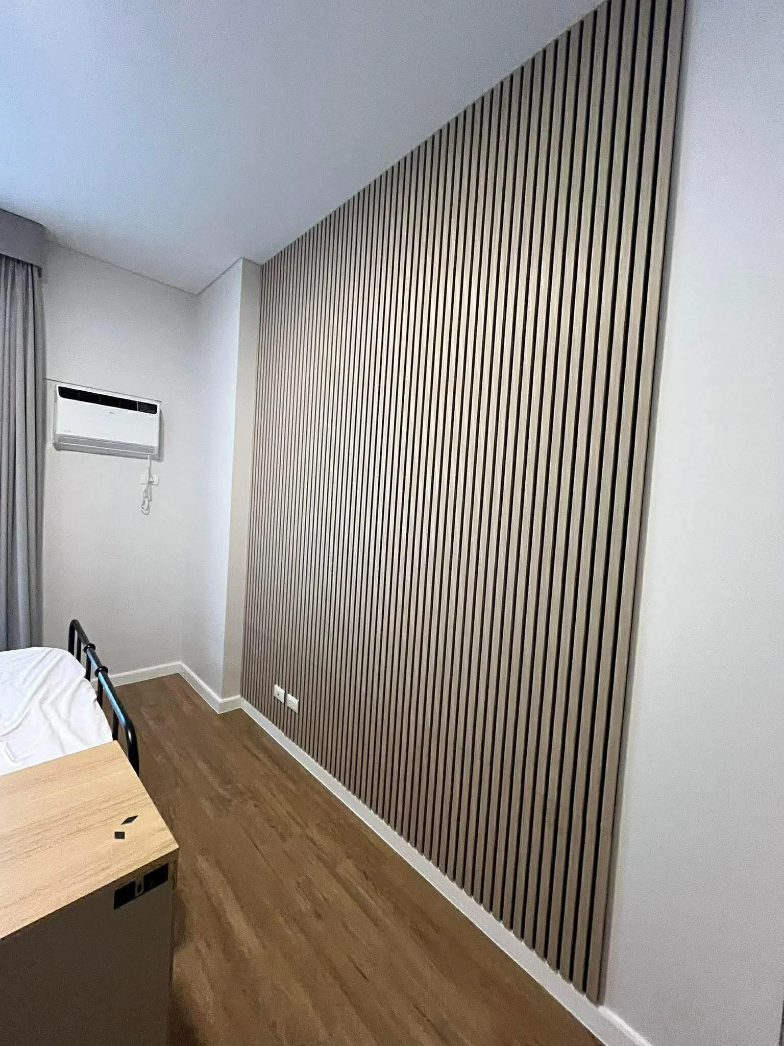 A bedroom corner featuring a wooden slat accent wall, a white air conditioning unit, a gray curtain, and a wooden nightstand.