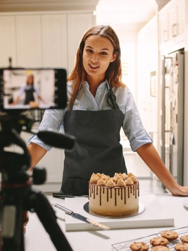 Female baker baking with a baked cake in front of her as she creates video content with a mobile phone on a tripod for social media
