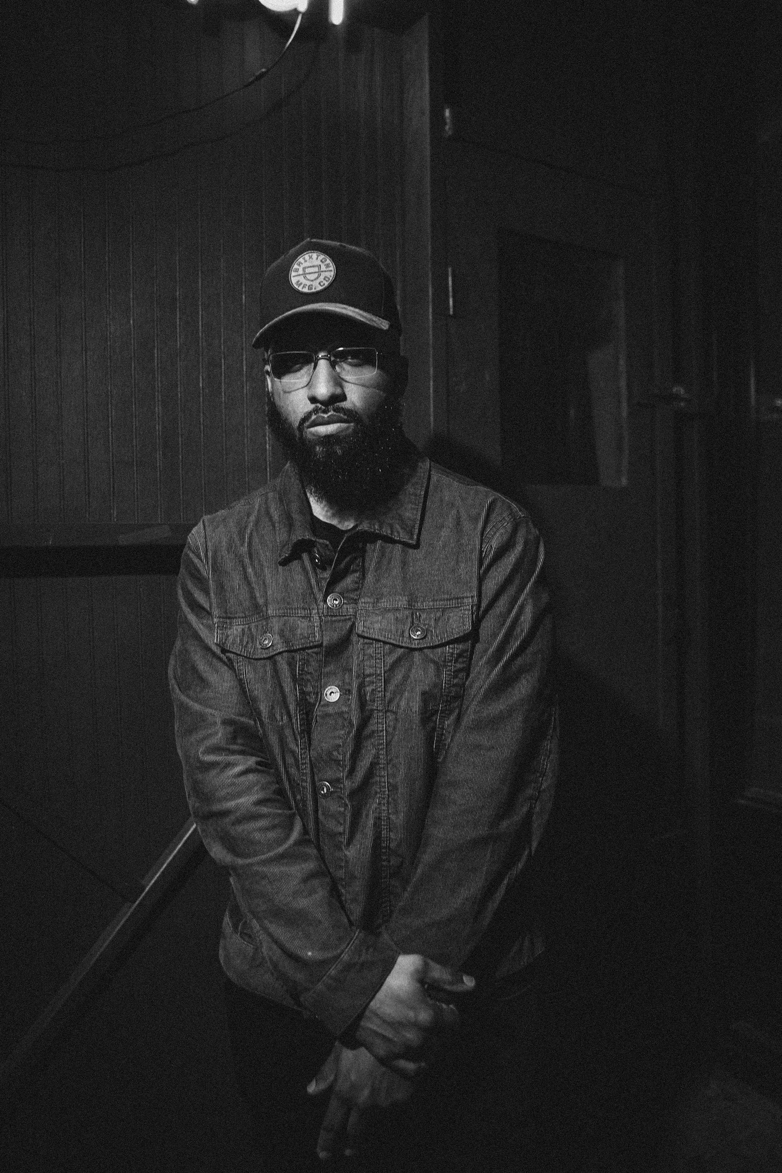 A man with a beard and glasses wearing a black cap and jacket, standing in front of a wooden wall. The image is in black and white.