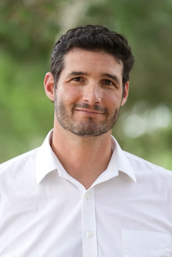 Portrait of a man with dark, curly hair and a beard, wearing a white dress shirt, standing outdoors with a blurred green background.