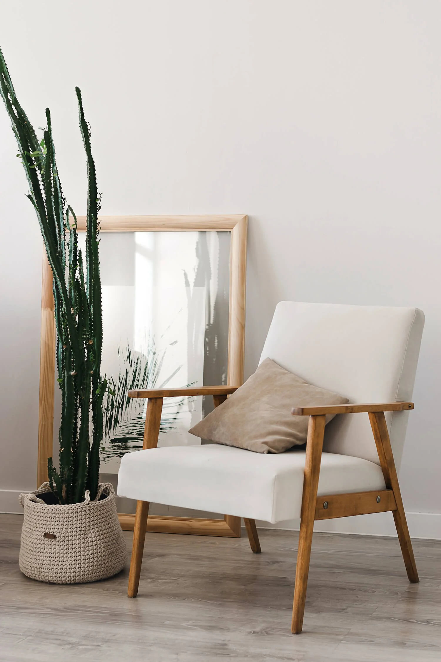 A cozy reading corner with a white upholstered armchair featuring wooden armrests and legs, a beige pillow resting on the chair, a tall cactus plant in a crocheted basket, and a framed mirror with a light wooden frame leaning against a plain white wall.