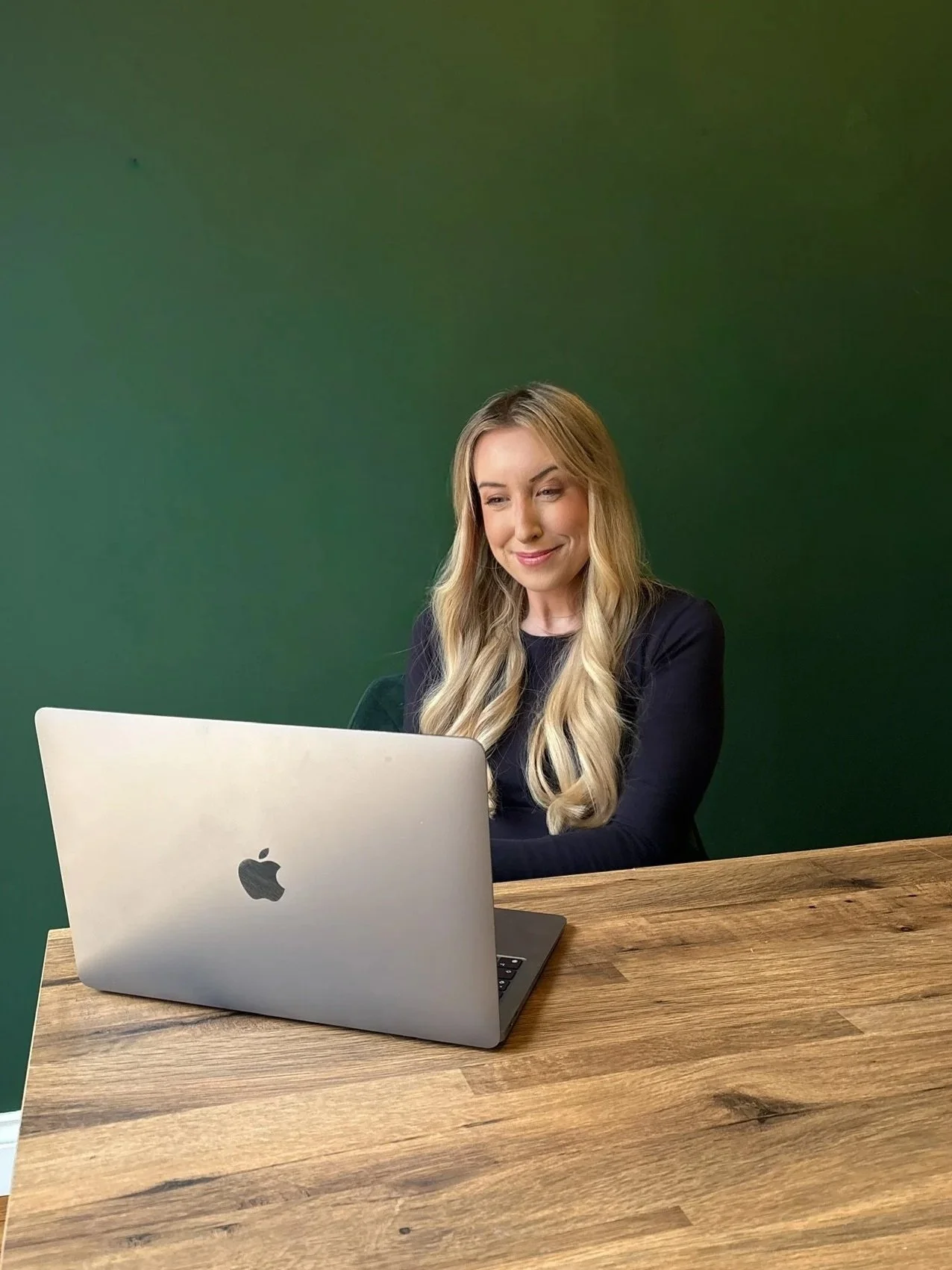 A woman with long blonde hair smiling while looking at a silver MacBook laptop on a wooden table, against a green wall background.