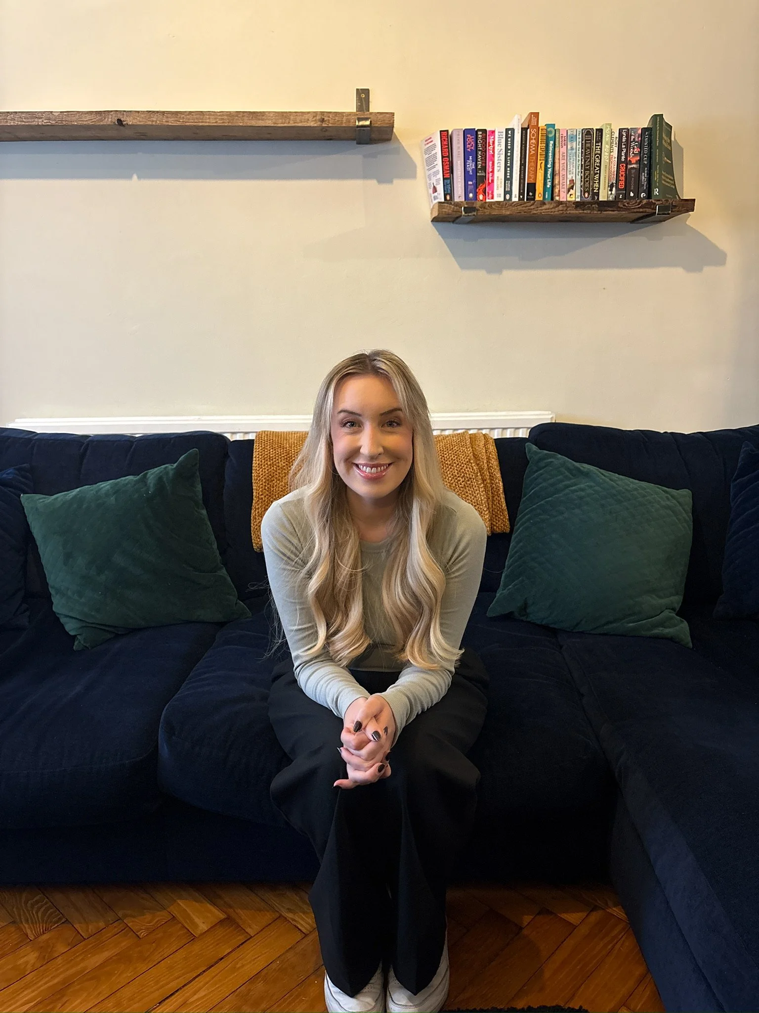A woman with long blonde hair sitting on a dark blue couch with green pillows, smiling at the camera. There are books on a wooden shelf on the wall behind her.