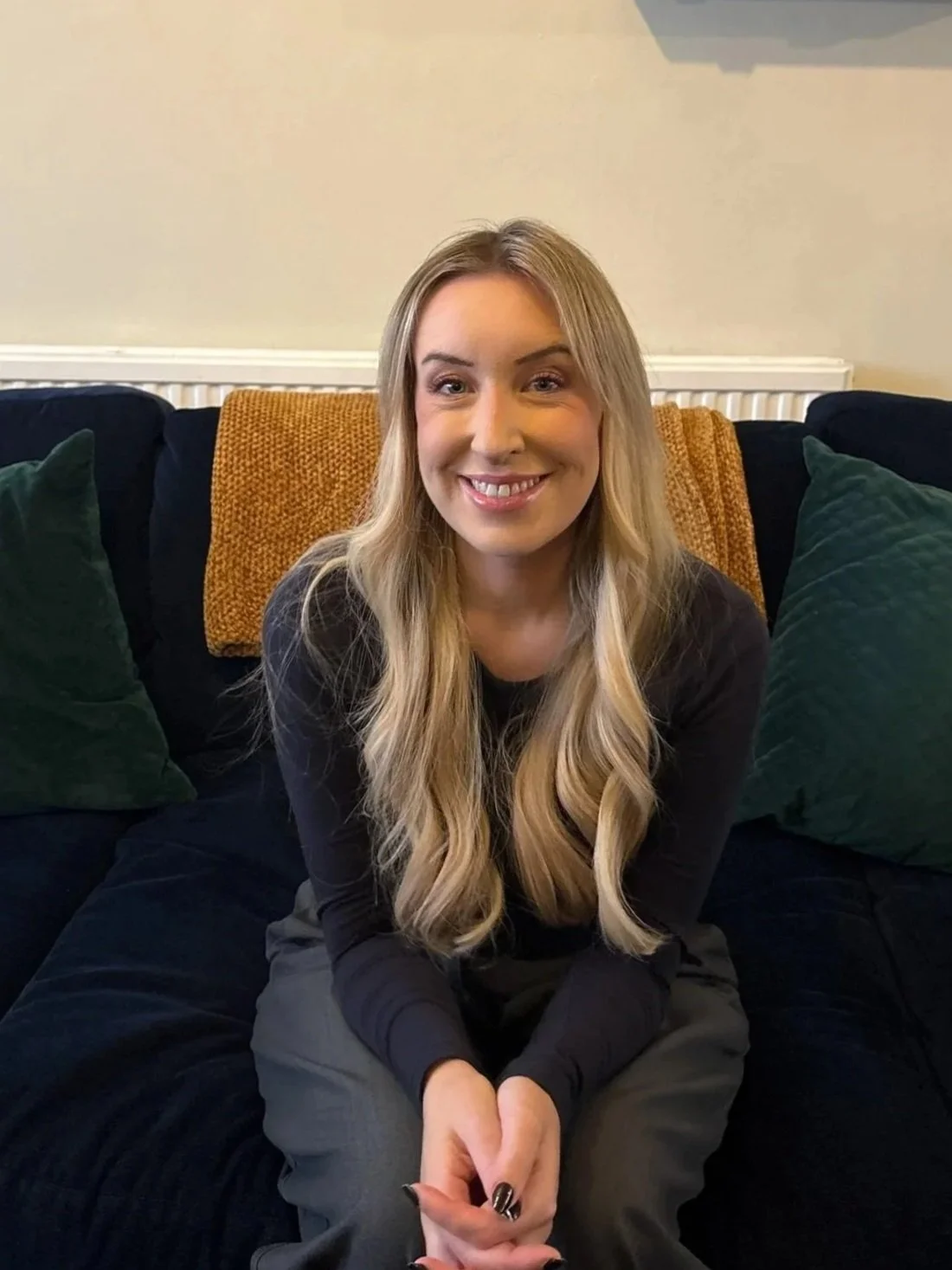 A young woman with long blonde hair, smiling, sitting on a dark-colored sofa with green pillows and a mustard-yellow blanket behind her.