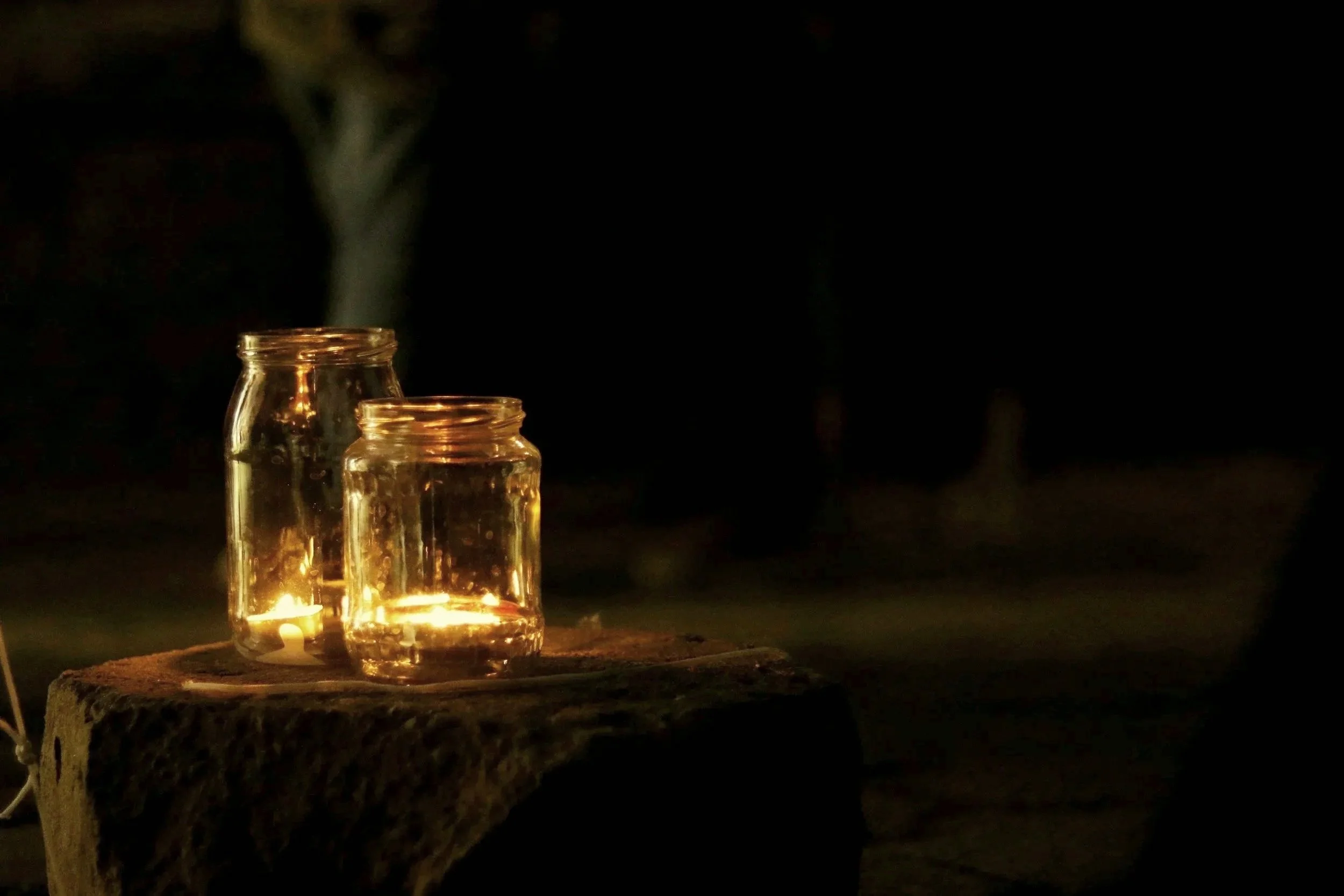 Two glass jars with candles inside placed on a wooden surface at night, with a dark background.