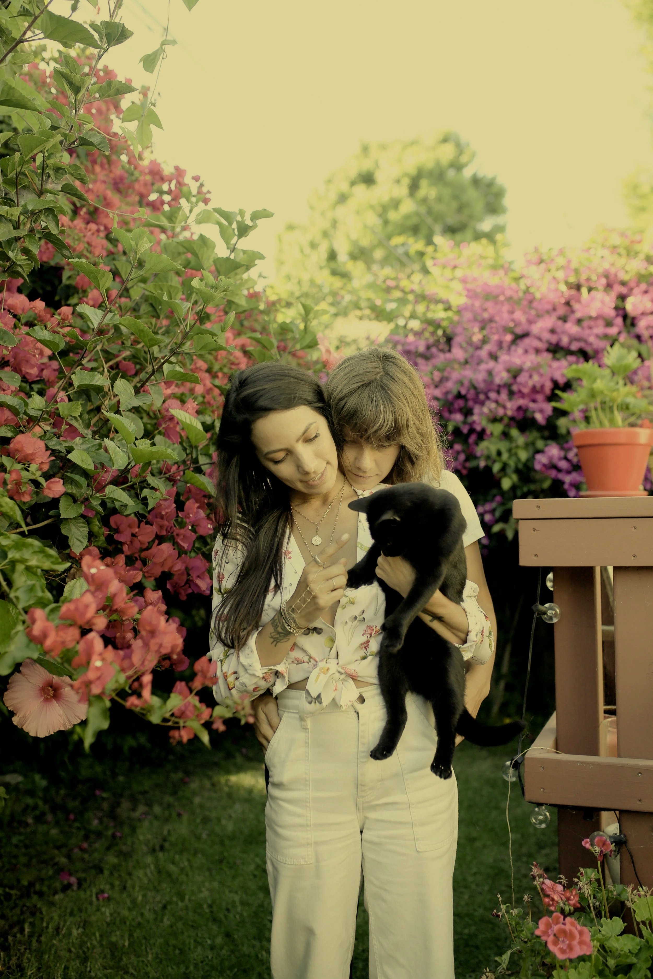 Two women standing in a garden with pink and purple flowers, one holding a black puppy, looking at it affectionately.