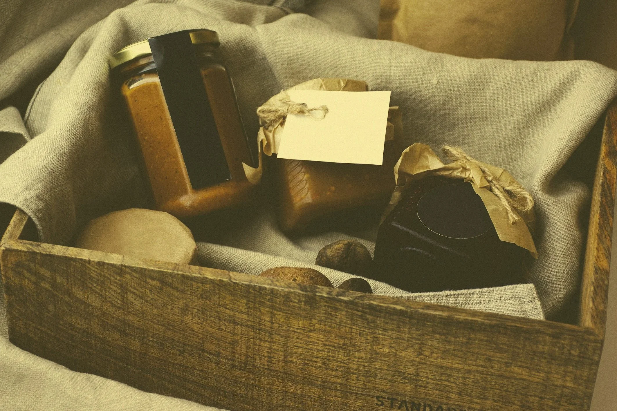 A wooden box containing jars of jam, a black jar, rocks, and a blank note, all lined with gray fabric.