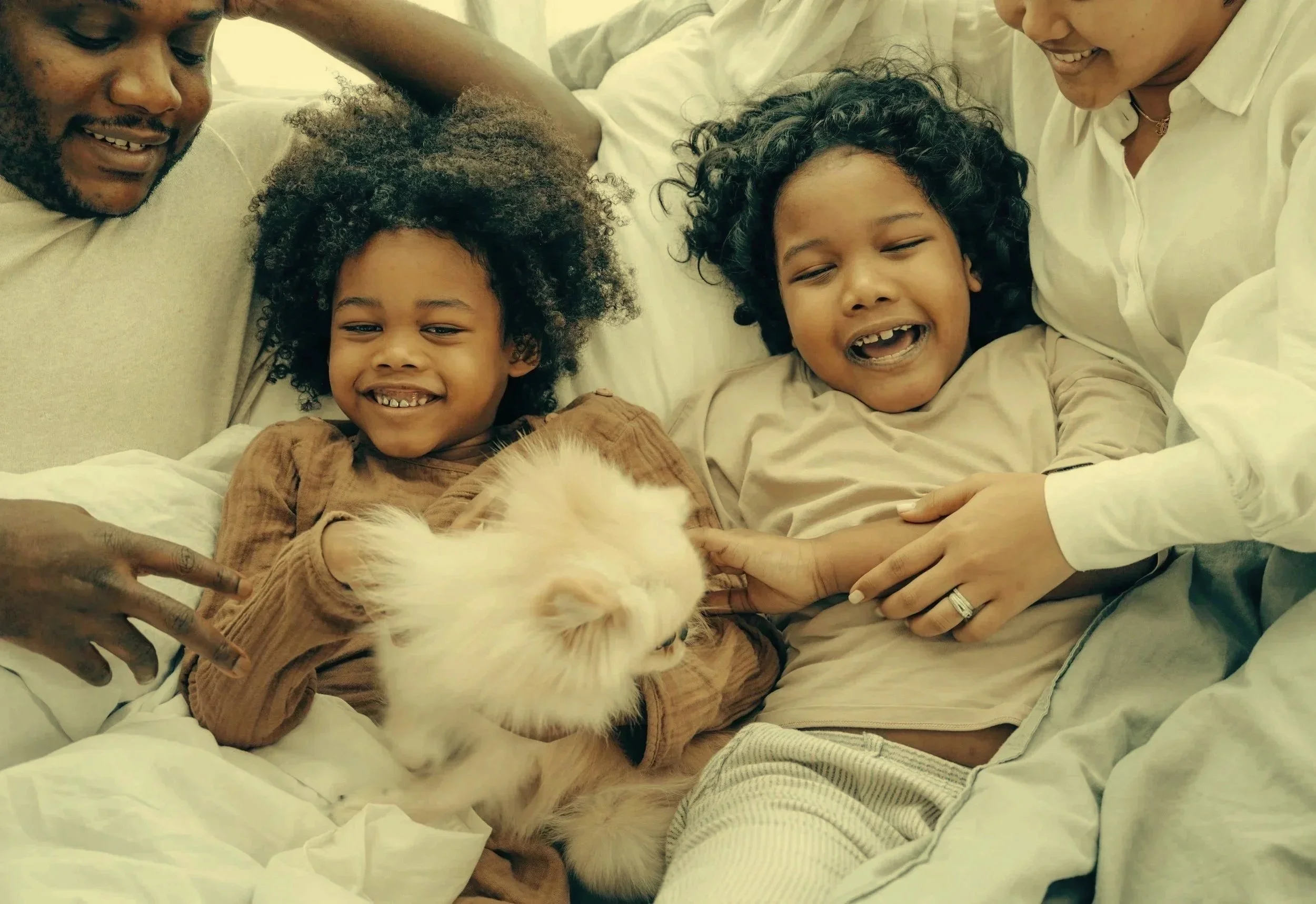 Family of four, including two children with curly hair and glasses, sitting on a bed with a small white dog, smiling and enjoying each other's company.