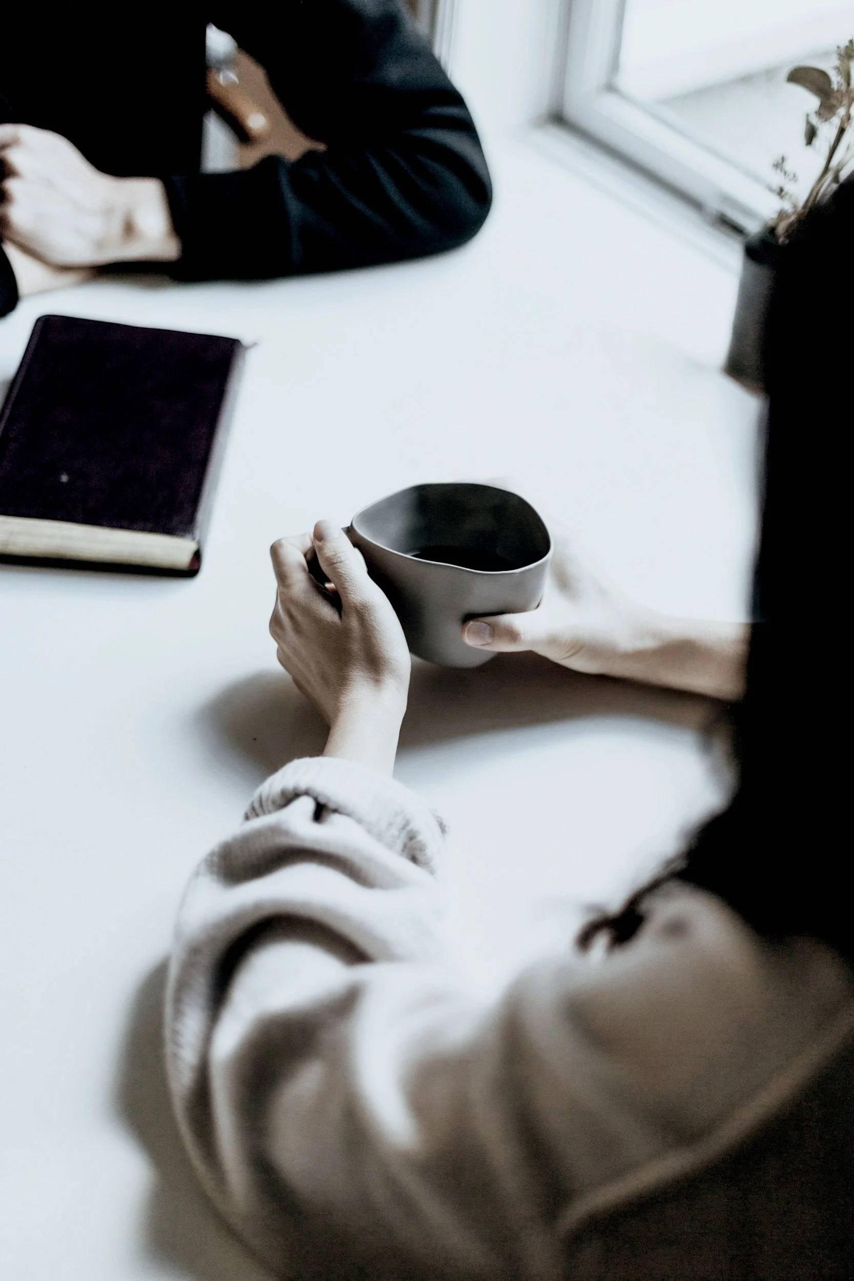 Person holding a ceramic mug with dark liquid, sitting at a white table next to a window.