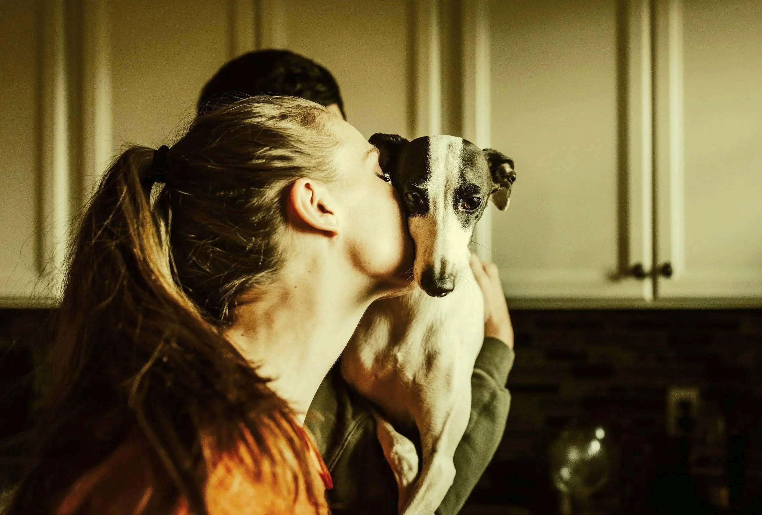 A woman is holding a whippet dog close to her face in a kitchen, with her eyes closed and kissing the dog, which looks calm and relaxed.