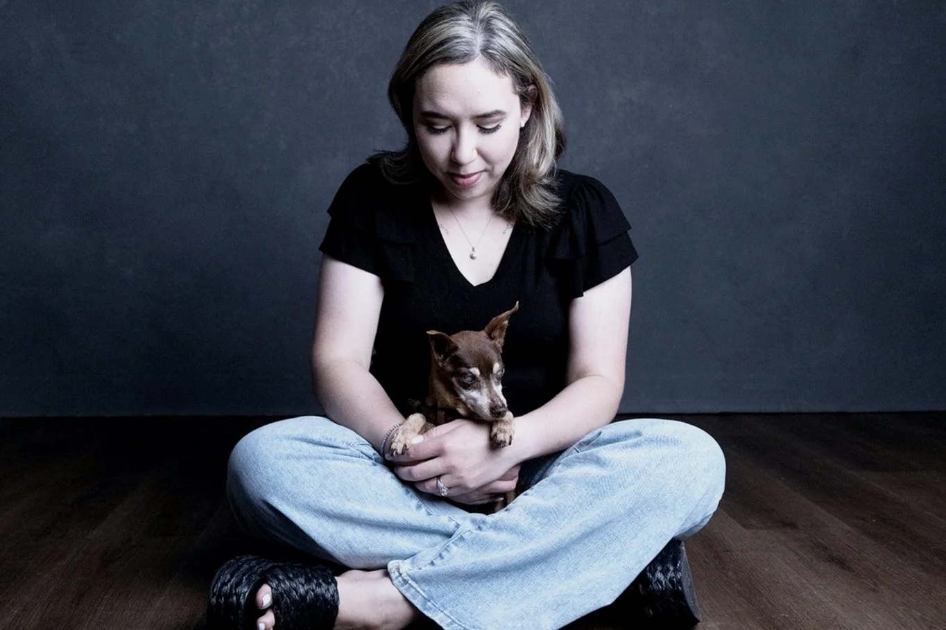 A woman sitting cross-legged on a wooden floor holding a small brown and tan puppy, with a dark background.