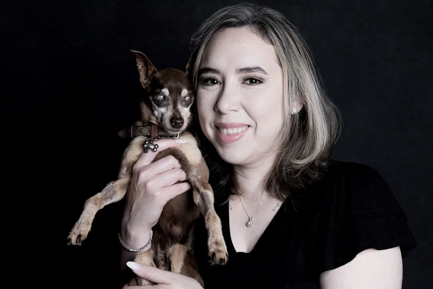 A woman with shoulder-length blonde hair, smiling, holding a small brown and black dog with blue eyes in front of a black background.