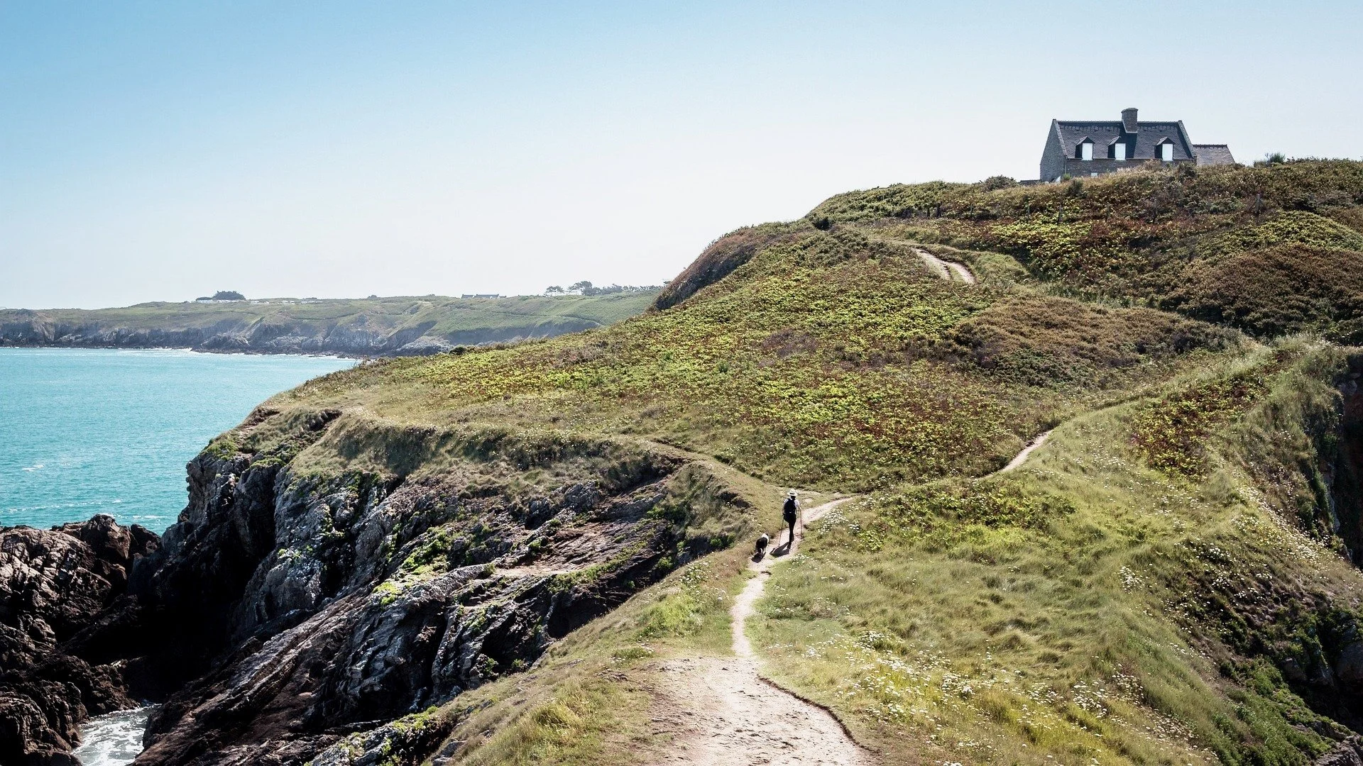 A person walking along a narrow trail on a green hillside overlooking the ocean, with a house at the top of the hill.