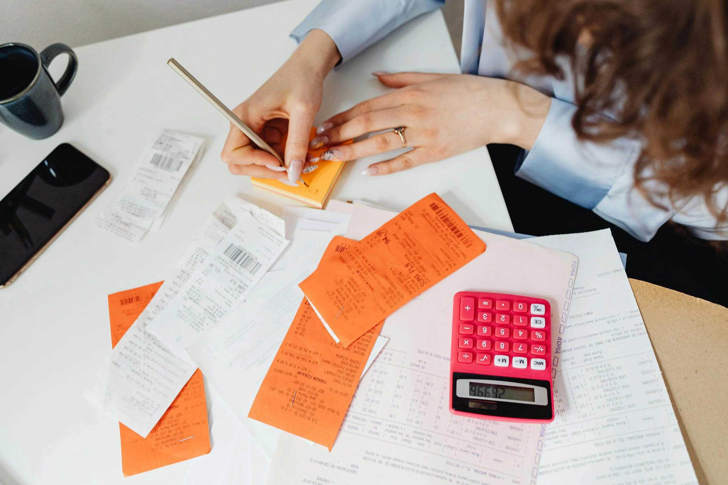A person is sitting at a desk with receipts, a calculator, a phone, and a notepad, working on finances or budgeting.