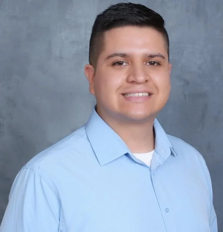 Portrait of a young man with short dark hair wearing a light blue button-up shirt smiling against a gray background.