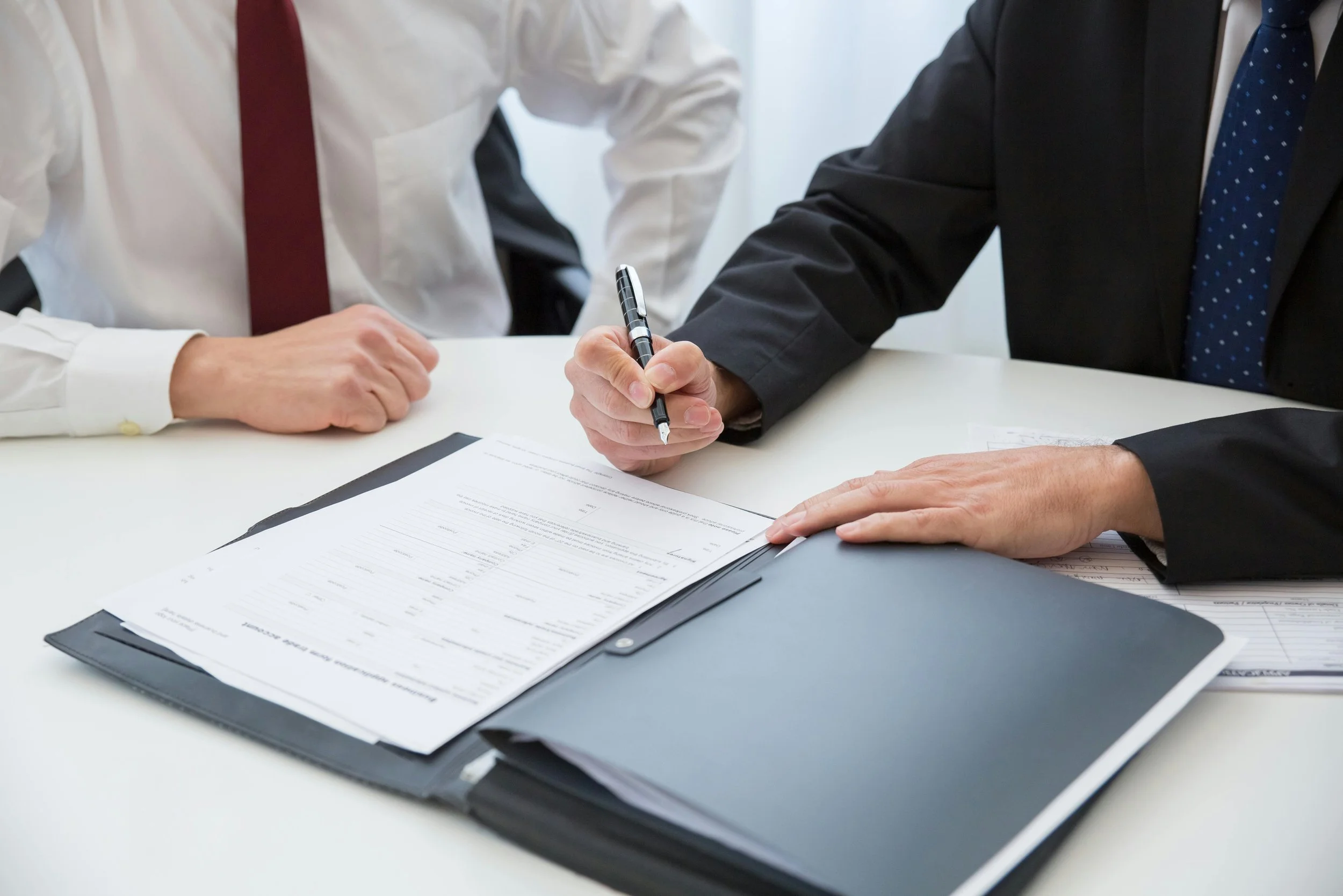 Two men in business attire sitting at a table reviewing documents, with one man pointing at paperwork with a pen.