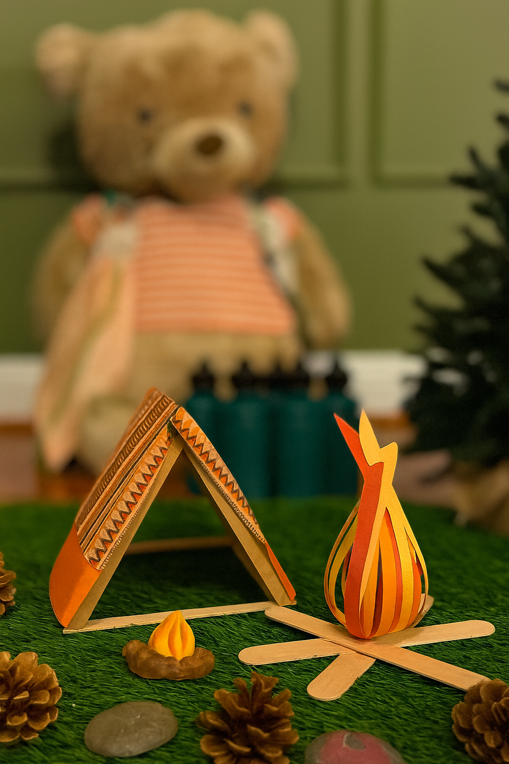 A miniature paper campfire, tent, and campfire with pinecones and stones on a green surface, with a blurred teddy bear in the background.