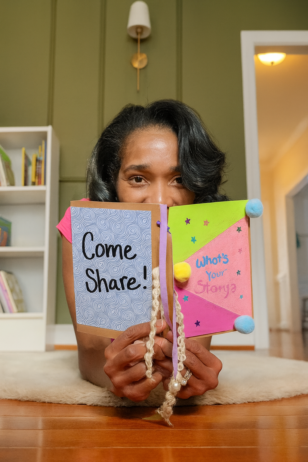 Woman lying on the floor holding a colorful handmade book that says 'Come Share! What's Your Story?' in a room with green walls, a white bookshelf, and a doorway in the background.