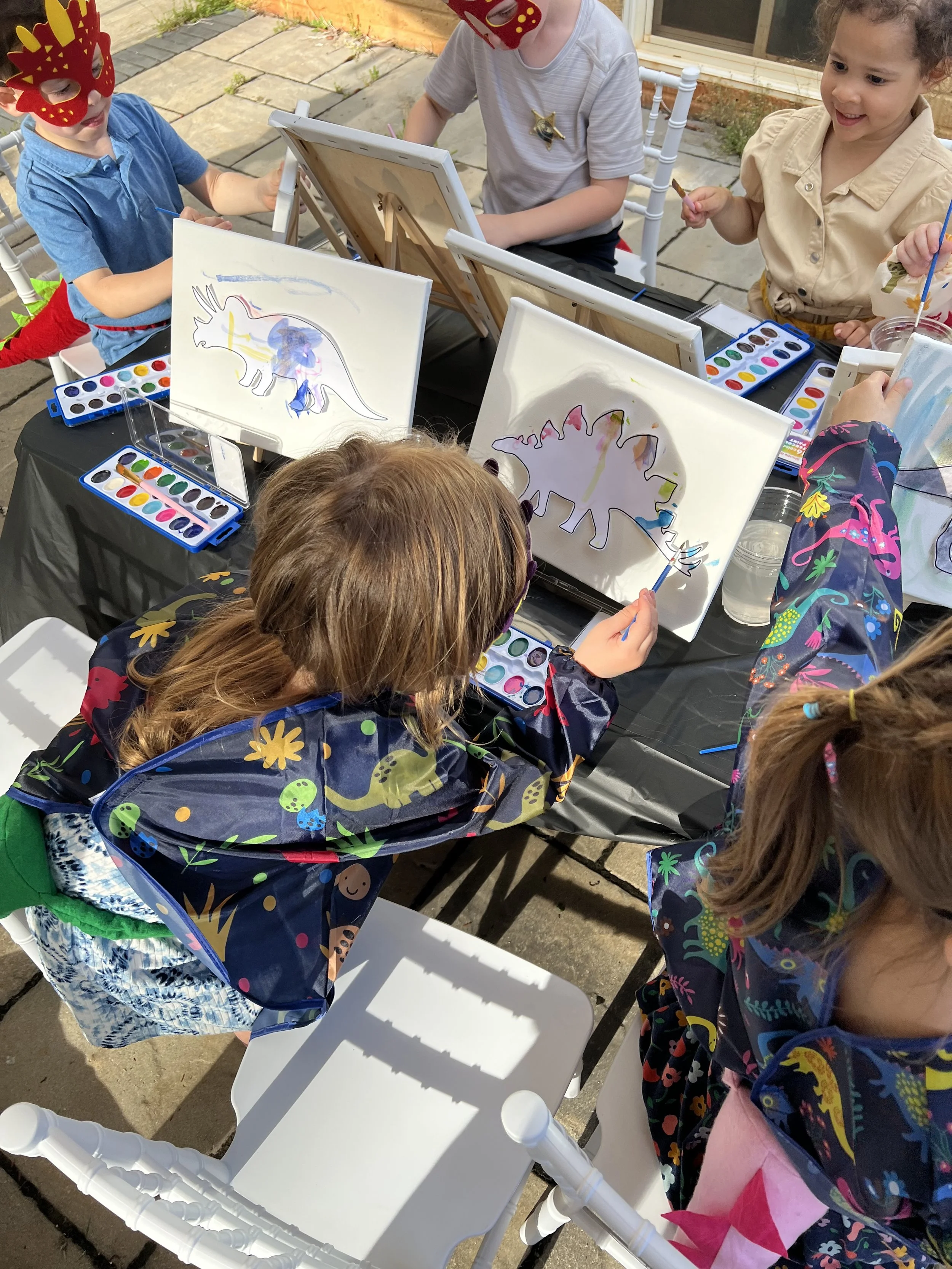 Five glass jars with sand, small rocks, and tiny green plants, each with a black plastic ring and a small black spoon inside. In the background, there are two toy dinosaurs, one green and one blue, and a small plant sculpture.