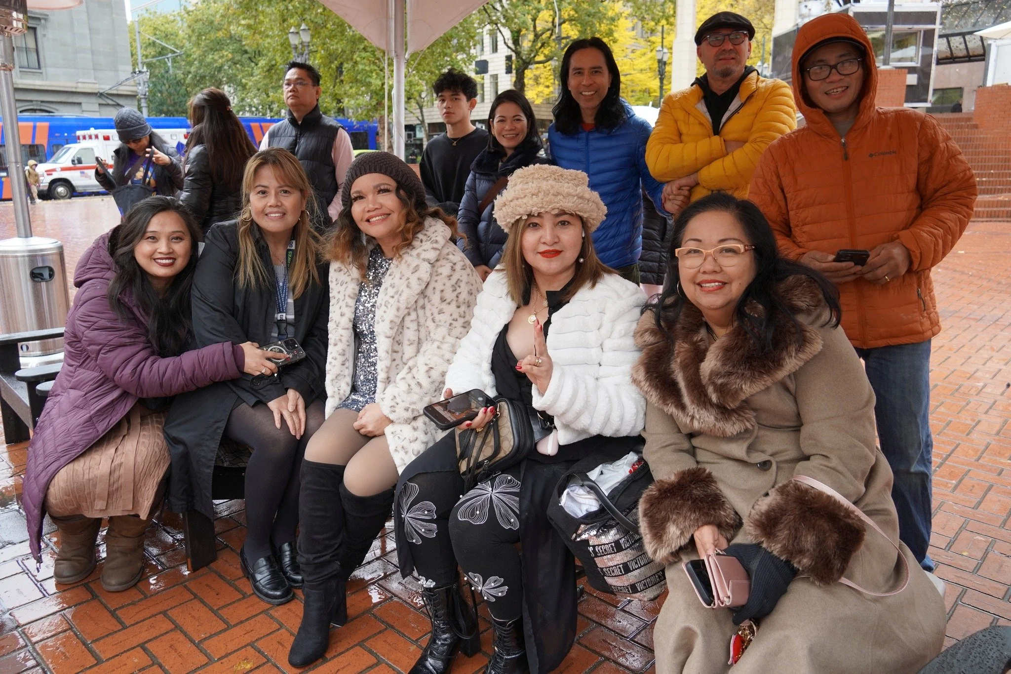 A group of ten people, five women and five men, gathered outdoors under a tent, posing and smiling for the photo with trees and buildings in the background. The women are sitting in the front row, wearing warm jackets and accessories, while the men s