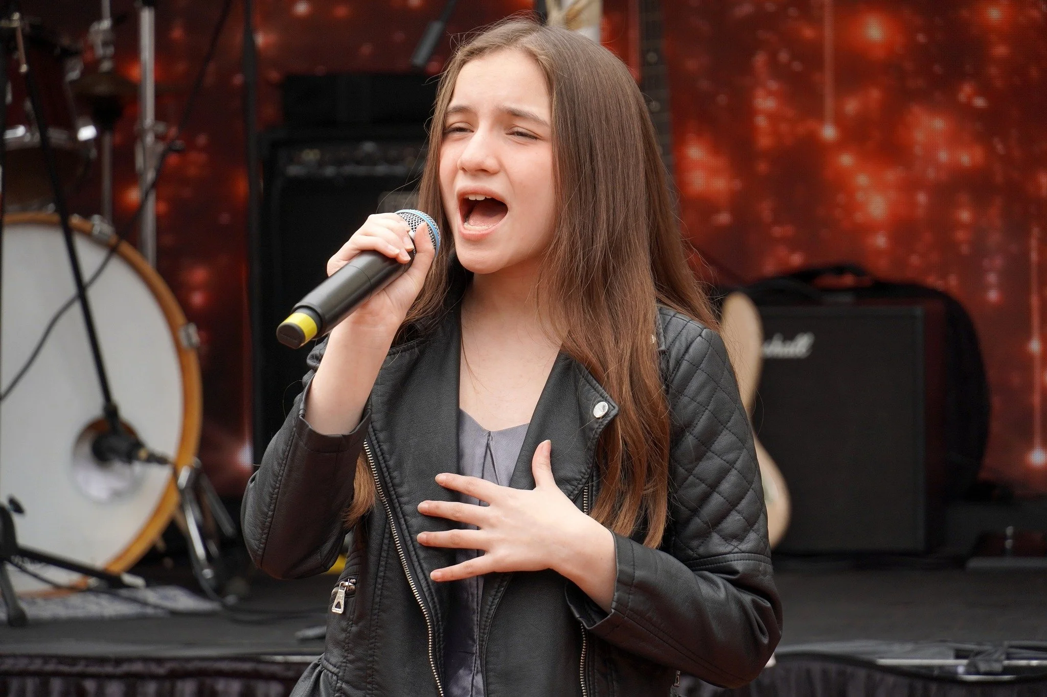 Young girl singing into a microphone, wearing a black leather jacket, with a musical stage setup in the background.