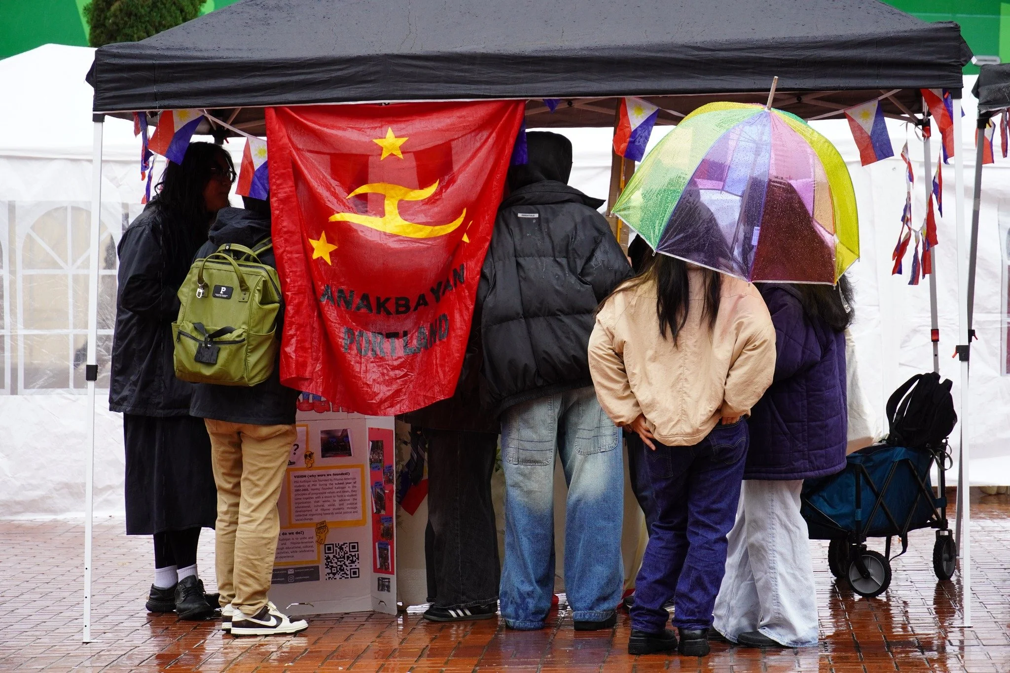 People gathered under a black tent with a red flag that reads 'Anakbayan Portland' and a yellow hammer and sickle symbol. Rain is falling, as evidenced by a colorful umbrella.