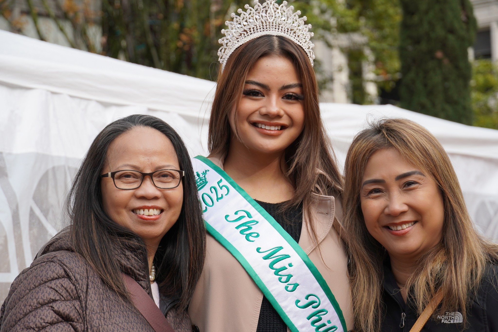 A young woman wearing a crown and sash that reads 2025 The Miss Philippines, standing between two older women outdoors, smiling at the camera.