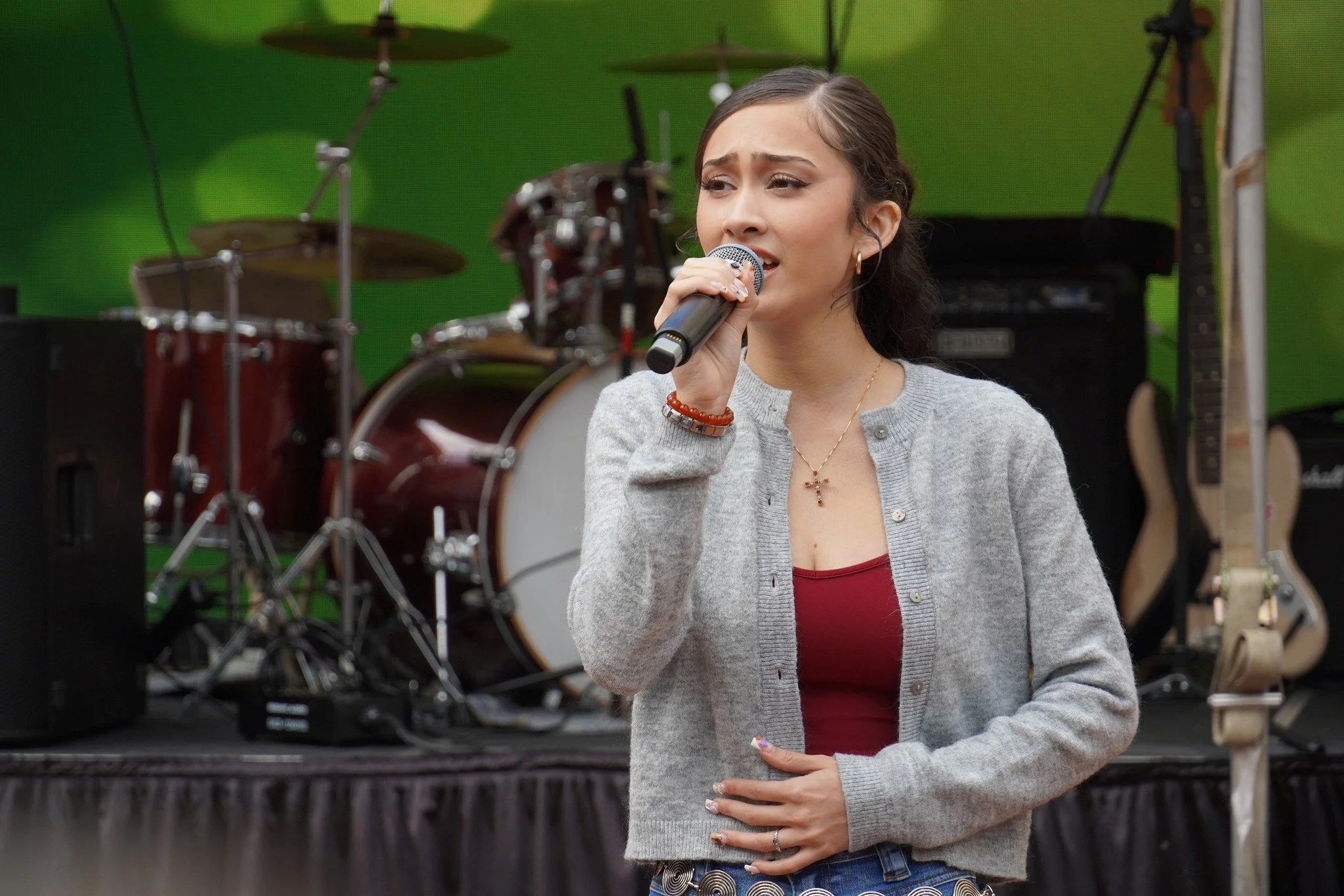 A young woman singing into a microphone on stage in front of musical instruments, with a green background.
