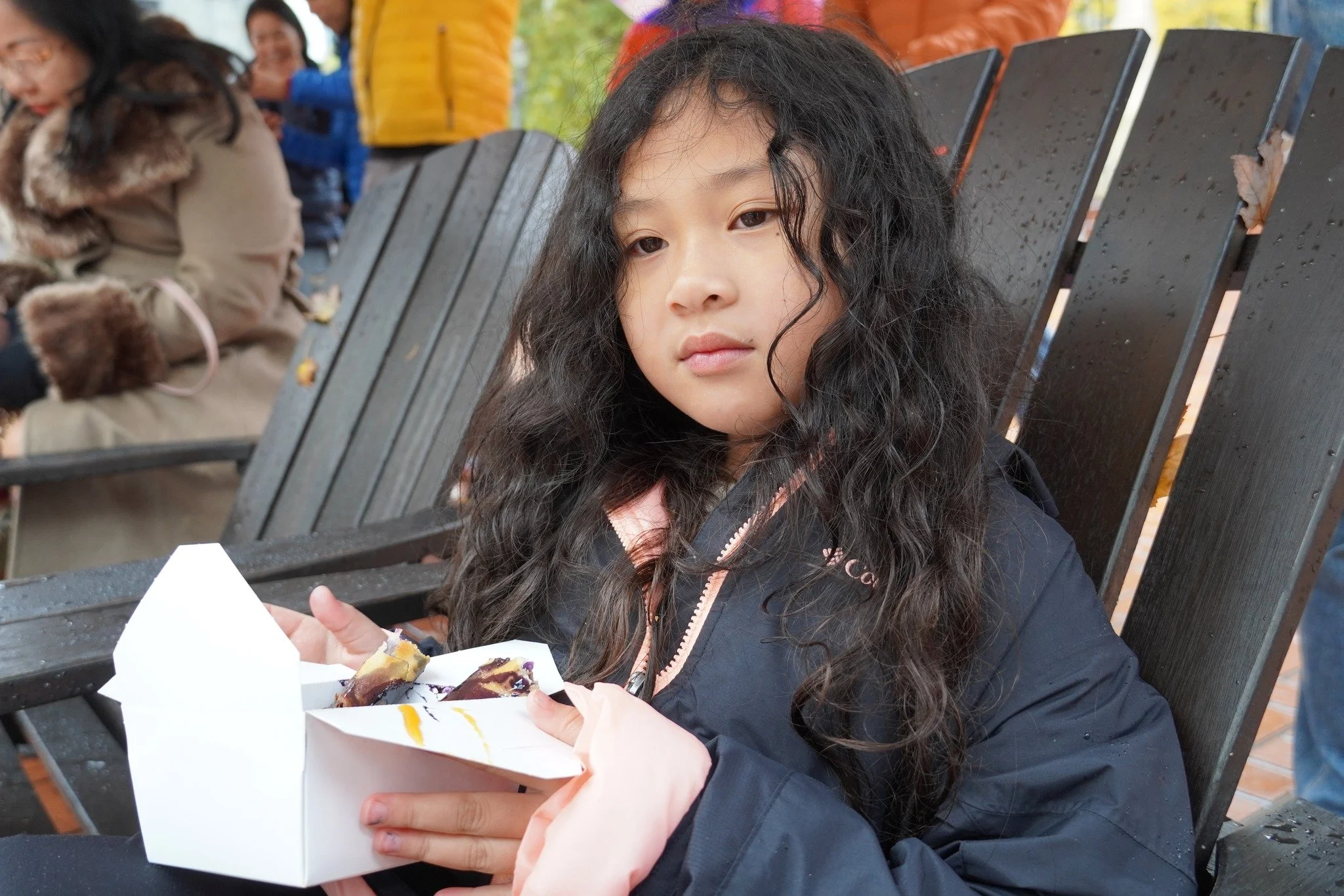 A young girl with long curly black hair sitting on a wooden bench, holding a box of dessert, outdoors with other people in the background.