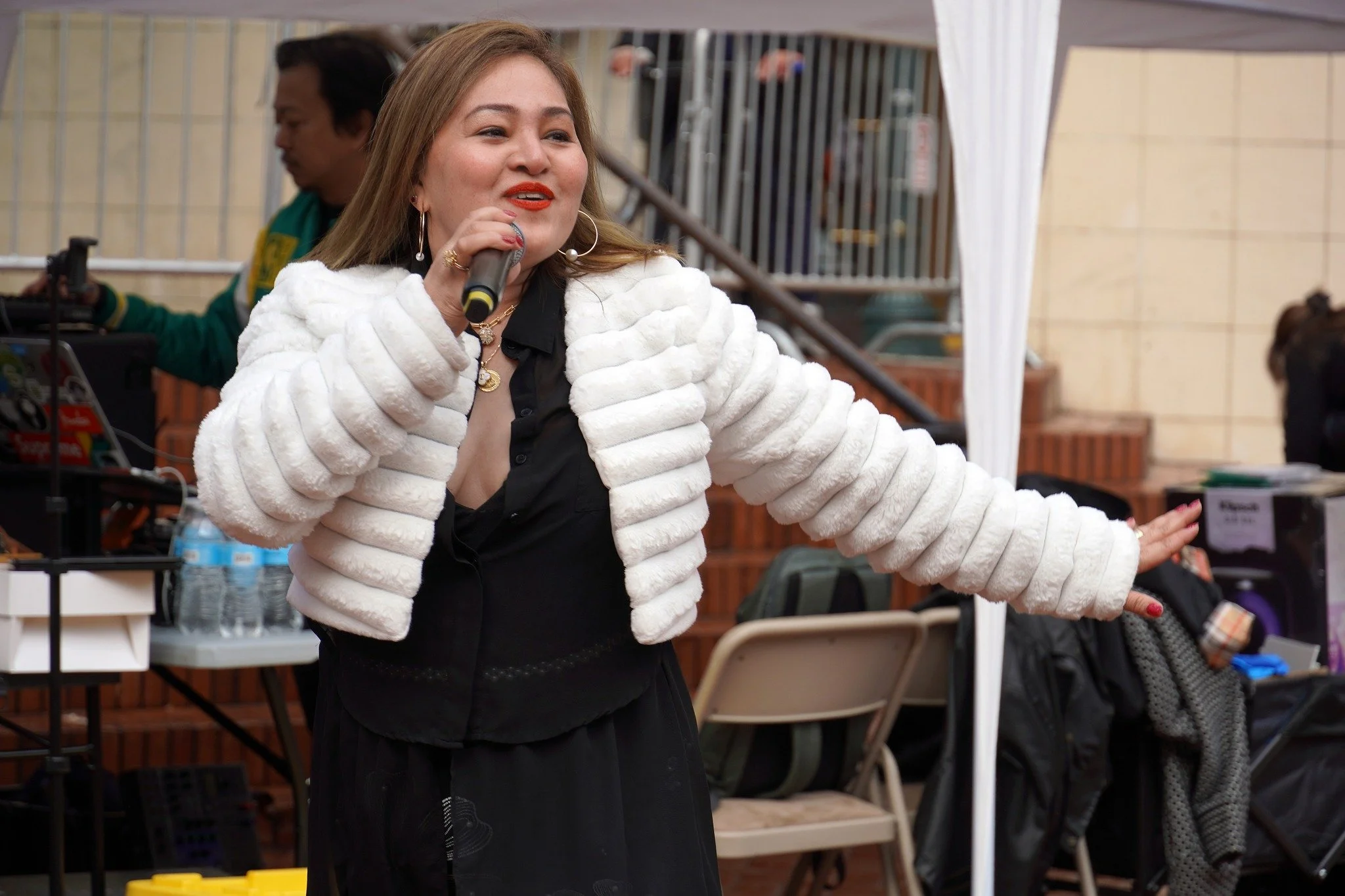Woman speaking into a microphone, wearing a white fur coat and black dress, standing in a venue with tables, chairs, and people in the background.