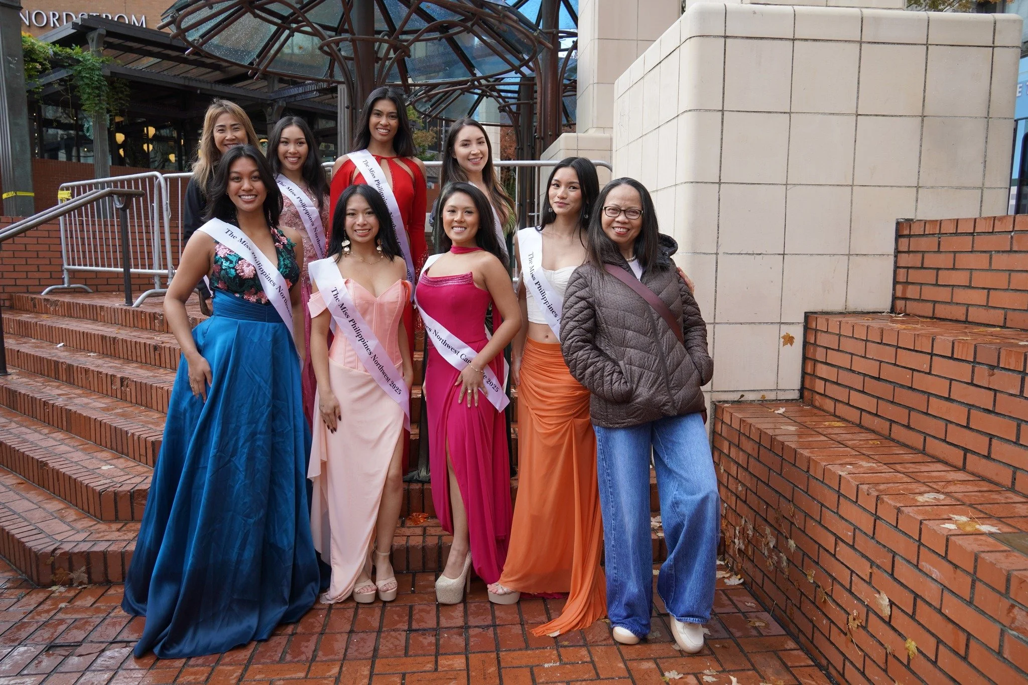 Group of women and one young girl standing on outdoor brick stairs, some wearing sashes and formal dresses, posing for a photo after a pageant event.