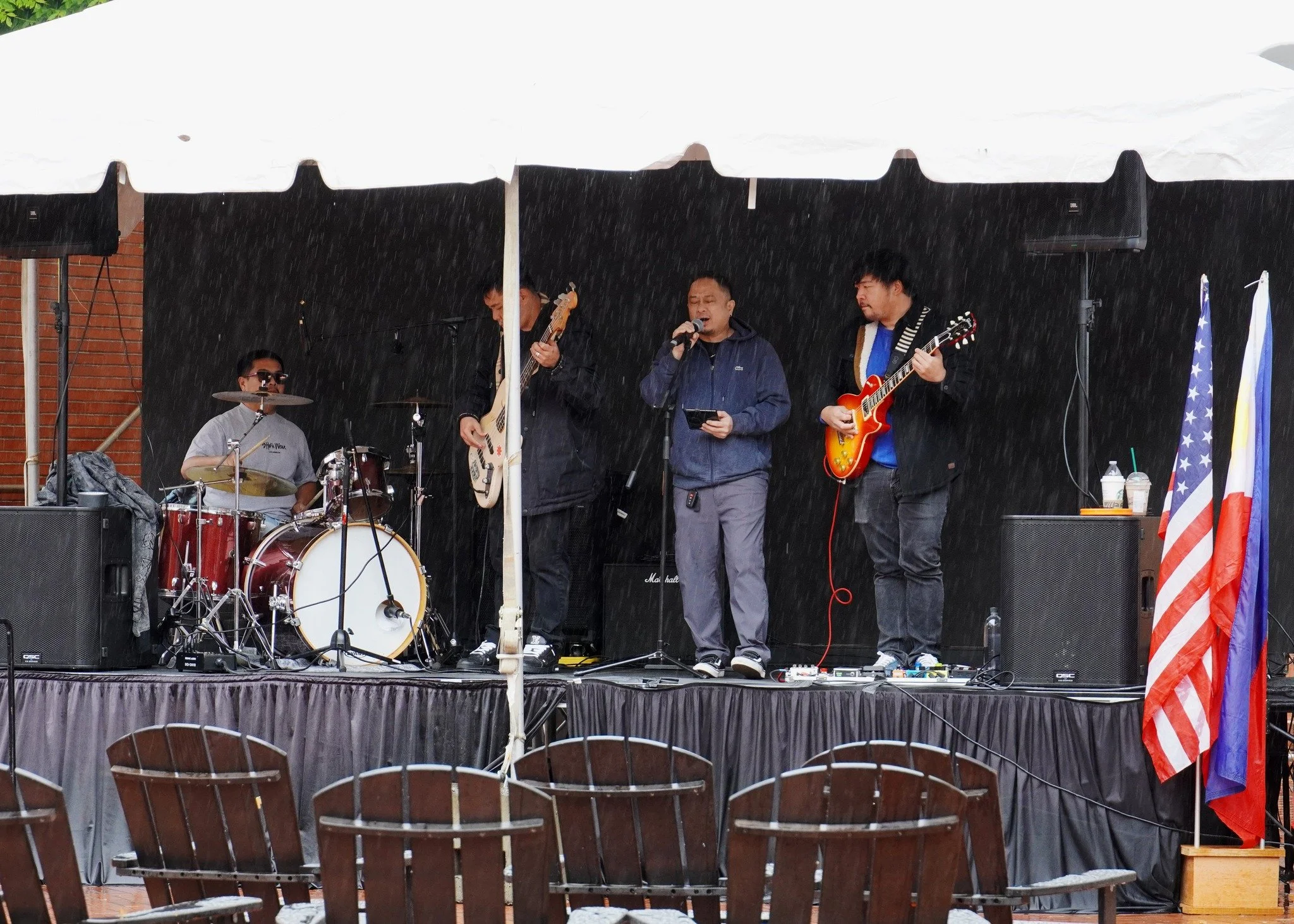 A band performing on an outdoor stage during rain, consisting of a drummer, two guitarists, and a vocalist.