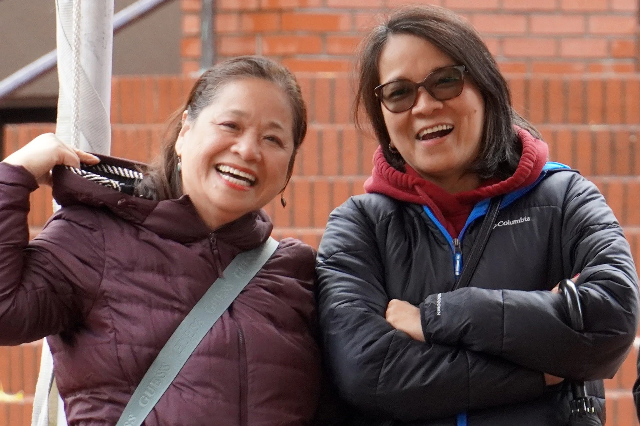Two women smiling outdoors, one wearing a maroon jacket and the other a black Columbia jacket and sunglasses.