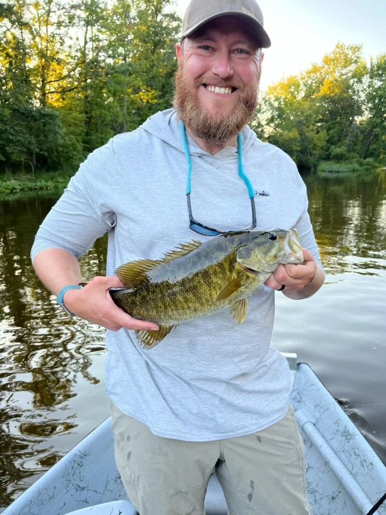 A man with a beard and gray cap smiling while holding a large fish on a boat near a lake, with green trees in the background.