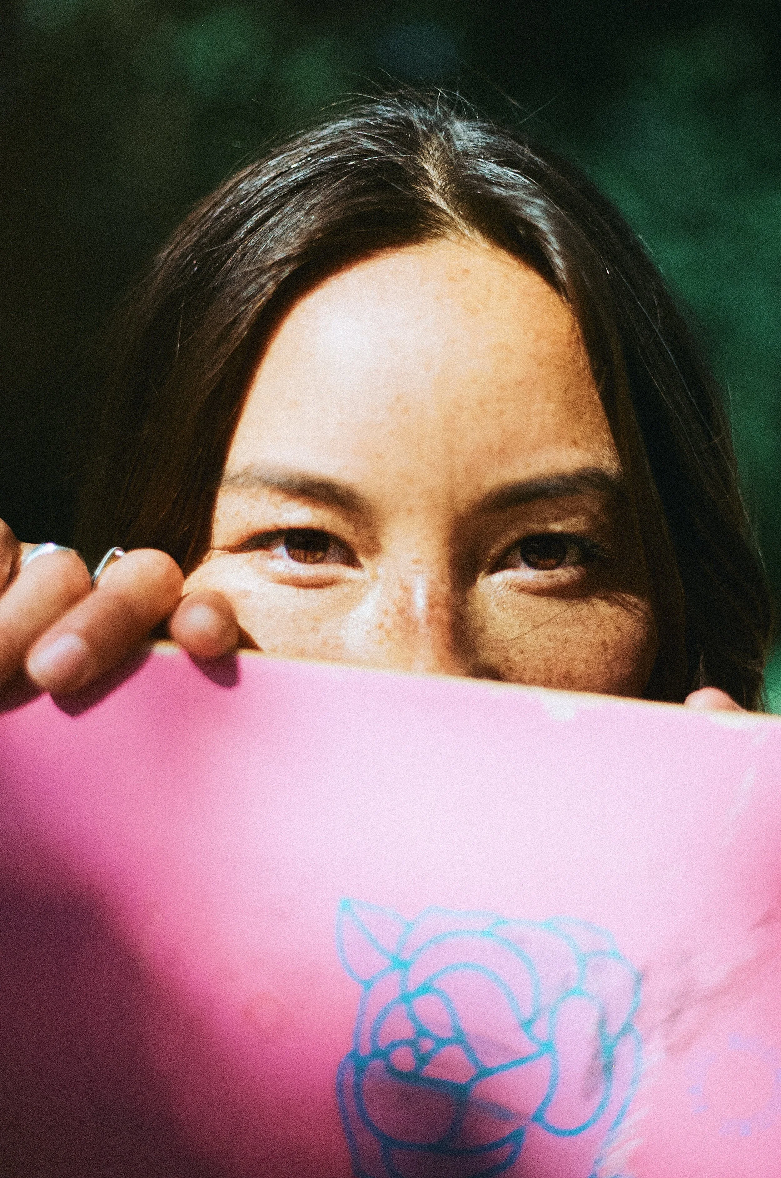 Close-up of a woman with brown hair holding a pink object in front of her face, partially covering it. She has a happy expression, with visible eyes and freckles on her face.